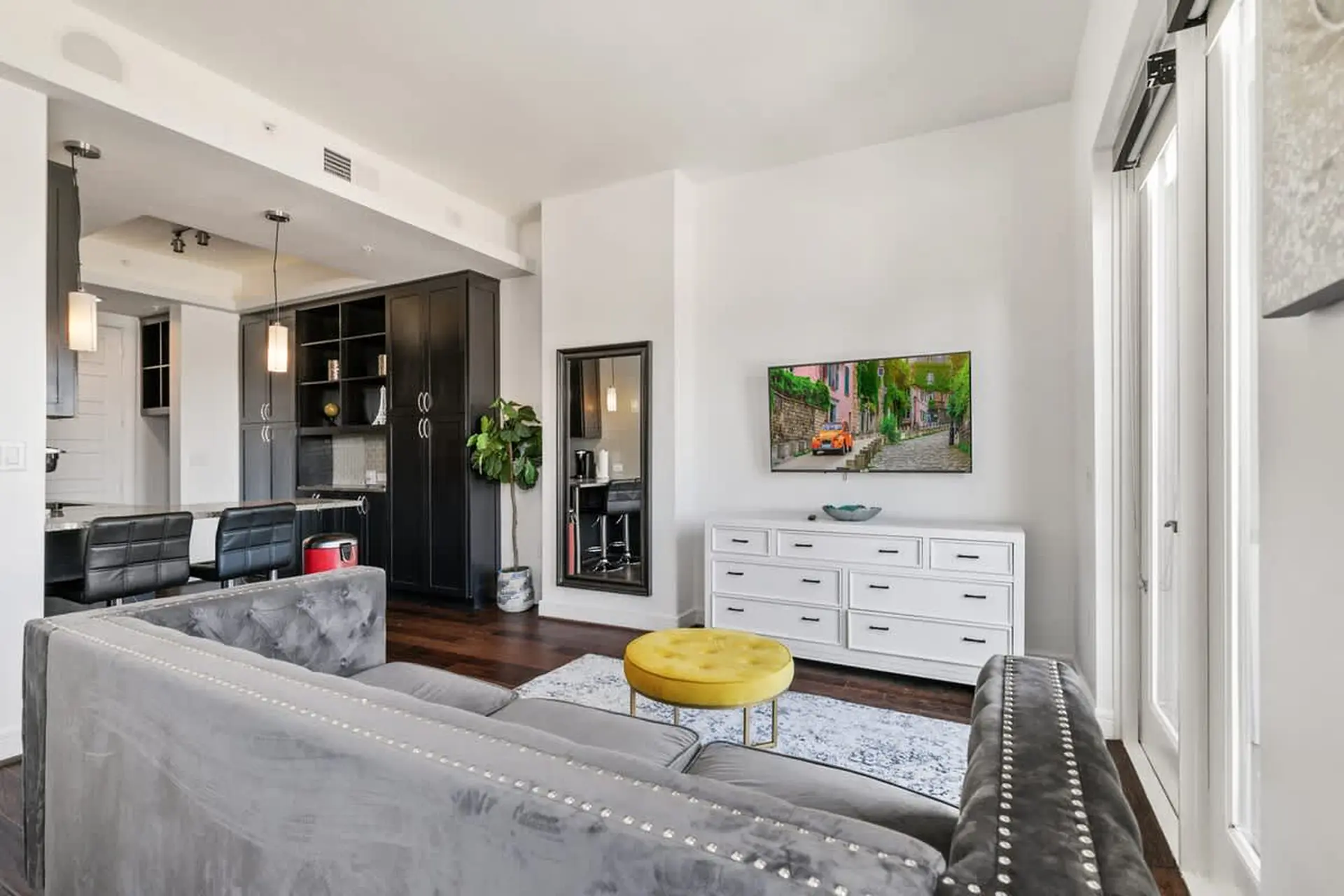 Living room with a gray sectional sofa, yellow ottoman, and a TV on a white dresser.