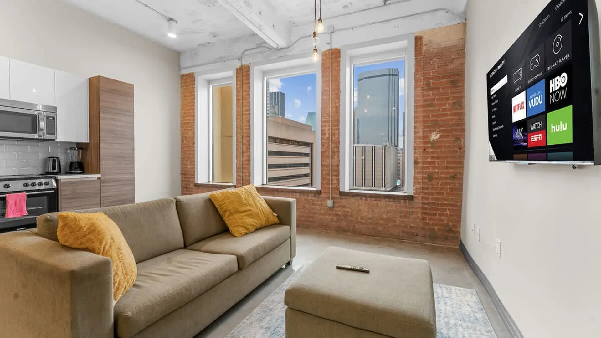 Loft apartment living room with a sectional sofa, ottoman, and large windows offering city views.