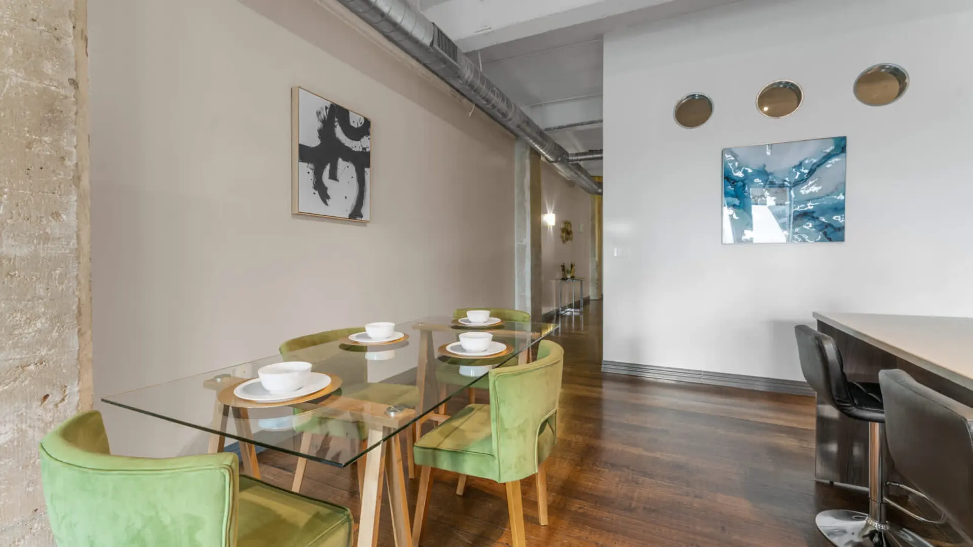 A dining table set with green chairs is in front of a wall with abstract art and round mirrors.