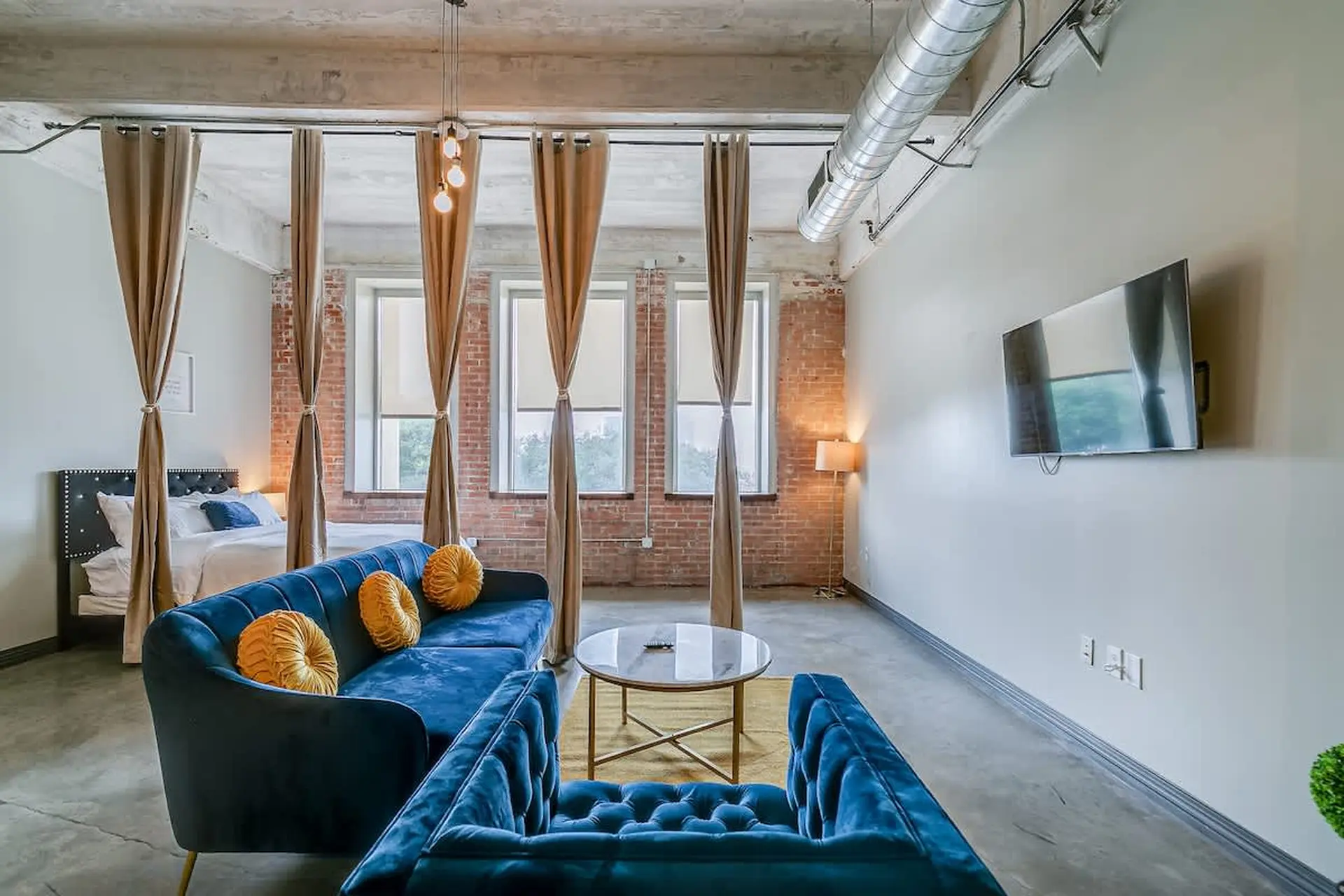 A blue velvet sofa and armchair face a coffee table, TV, and bed in a loft apartment.