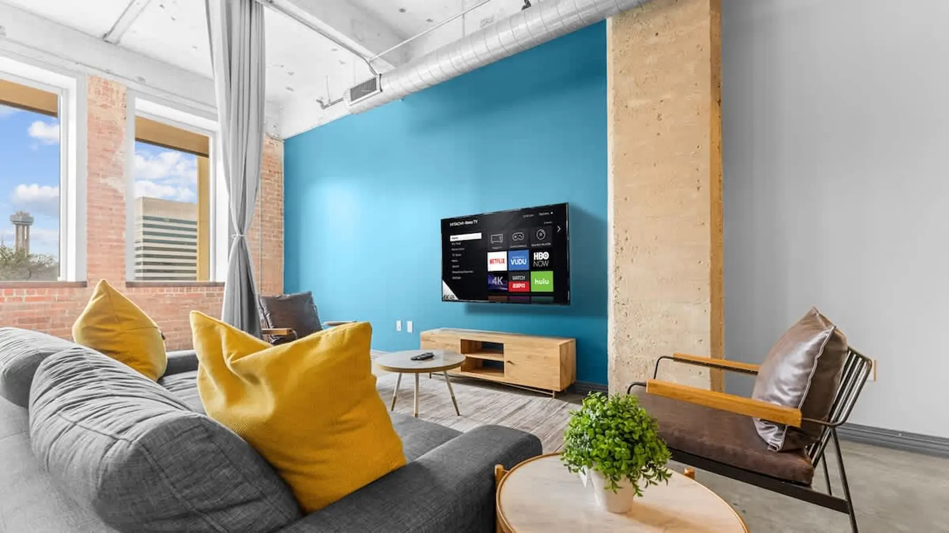 A modern living room with a gray sofa, yellow pillows, and a TV mounted on a blue wall.