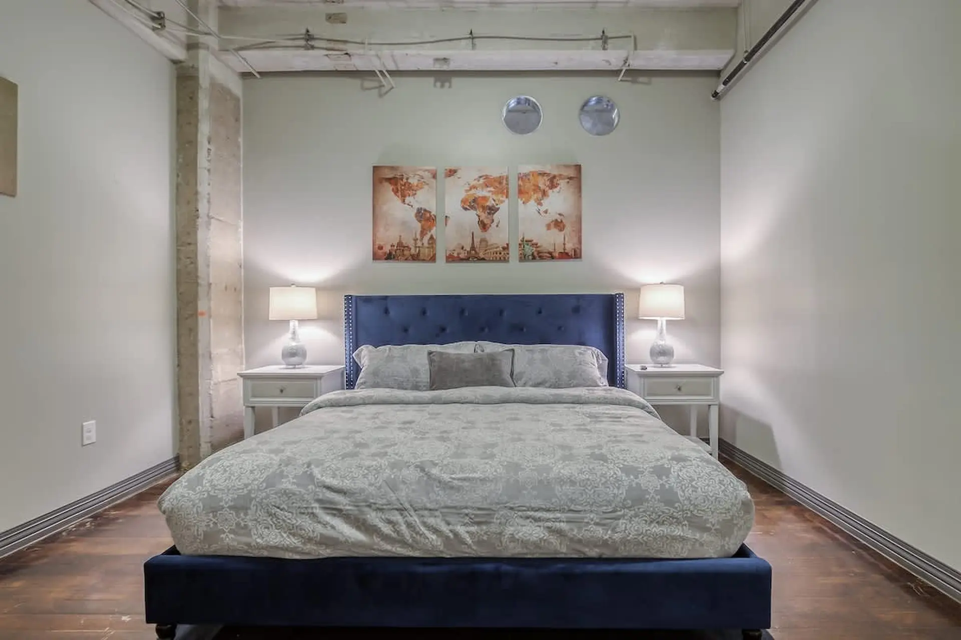A bedroom with a blue tufted headboard, world map art, and matching nightstands with lamps.