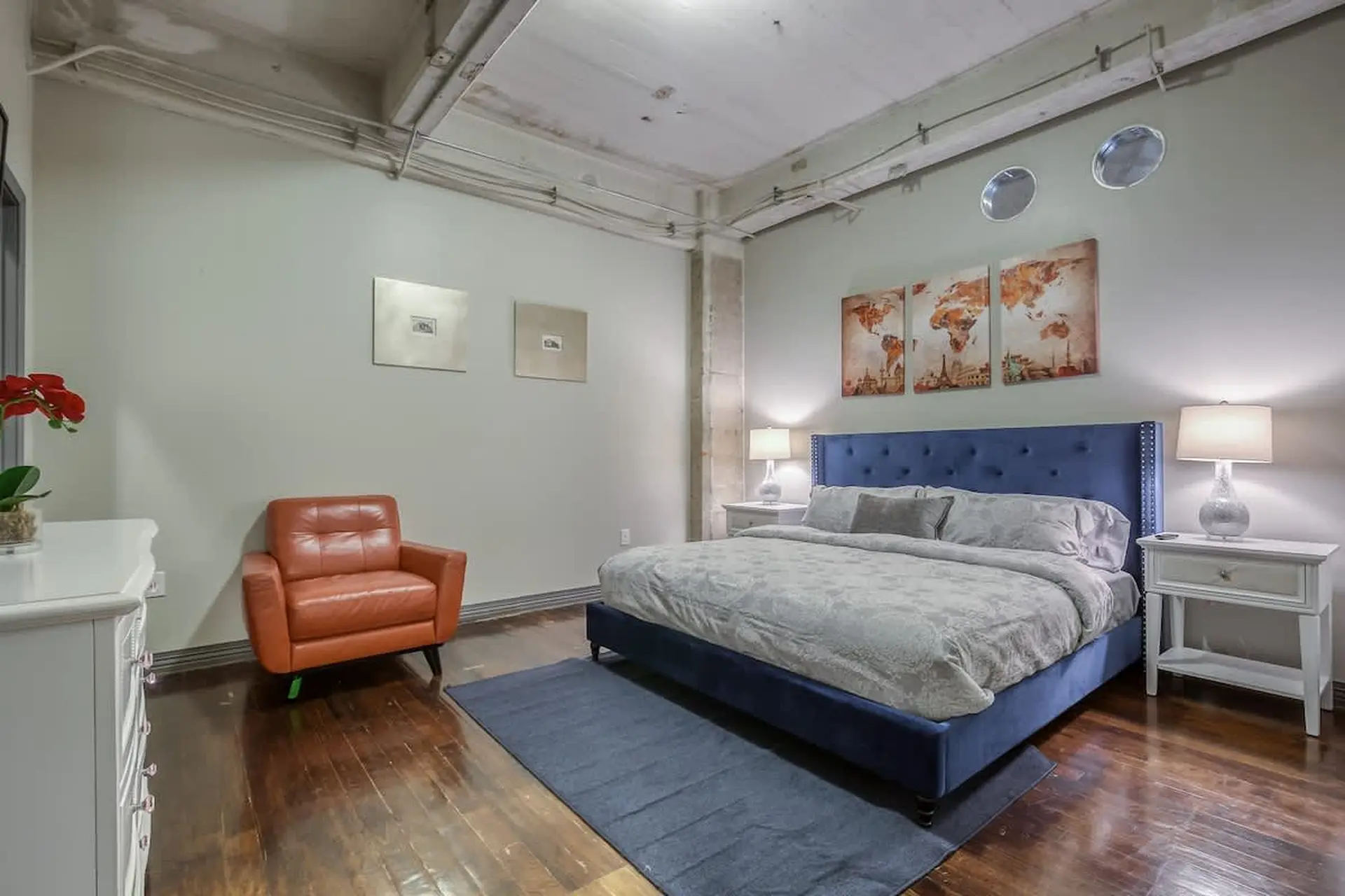 Bedroom with a blue tufted bed, orange armchair, and cityscape art.