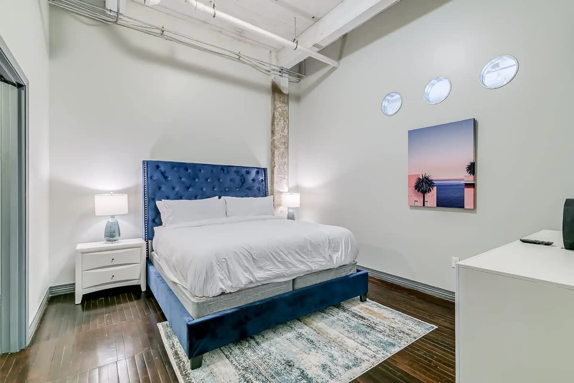 Modern bedroom with a king-size bed, blue headboard, nightstand, and artwork on the wall.