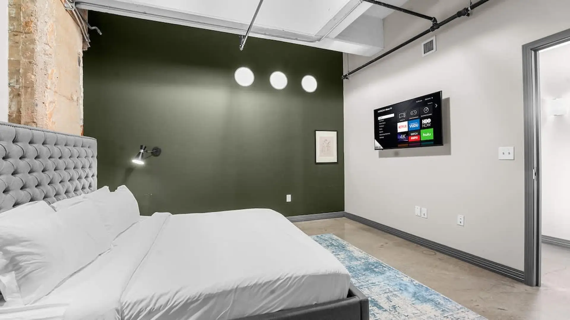 A bed with white sheets and a gray headboard faces a wall with a TV.