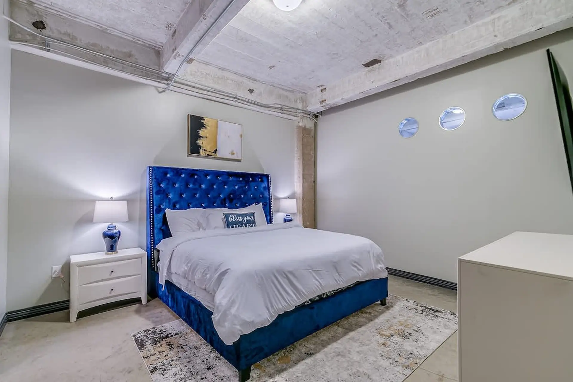 A bedroom with a blue velvet bed, white nightstand, and abstract art.