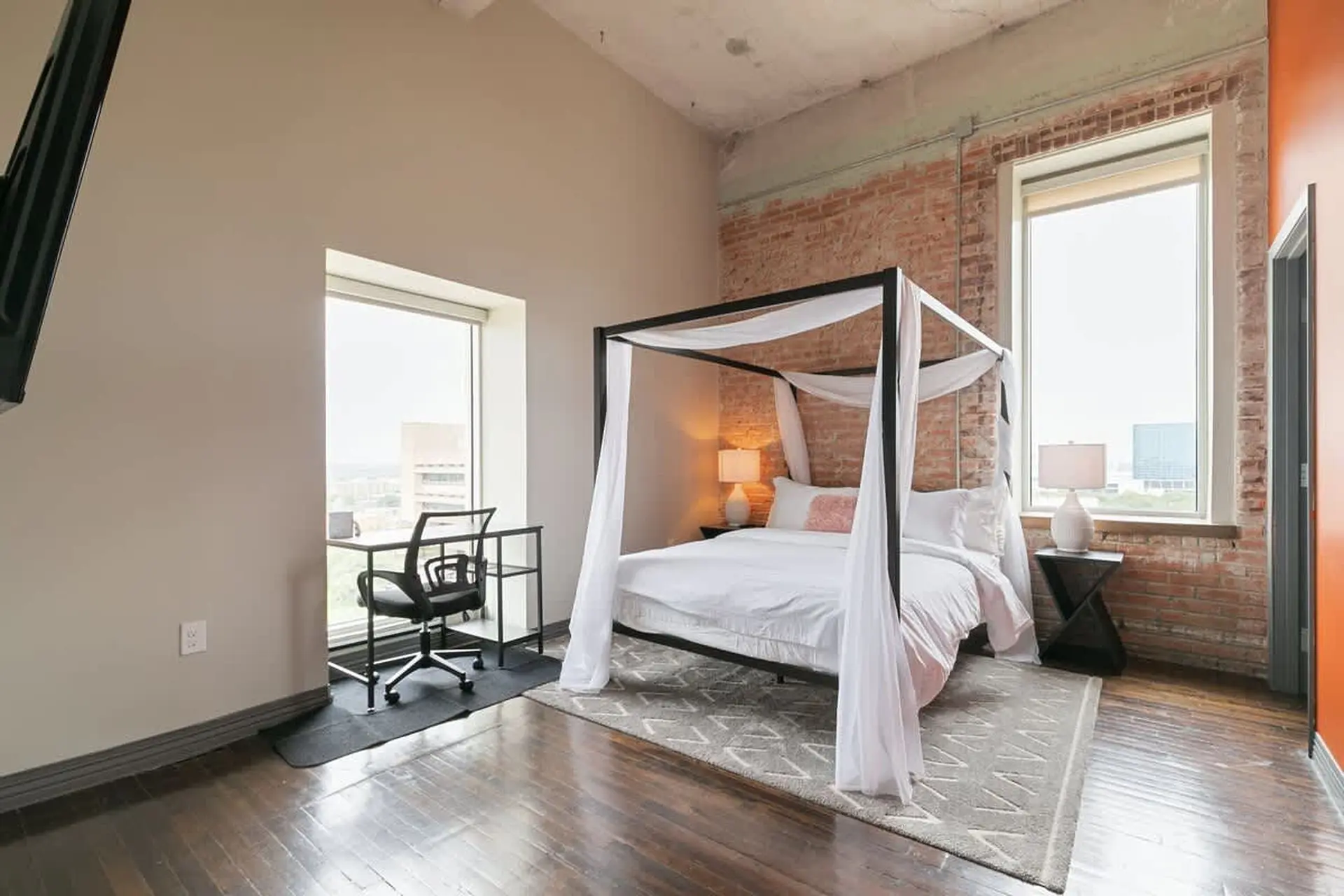 A white canopy bed with white curtains stands in a bedroom with a brick wall and large windows.