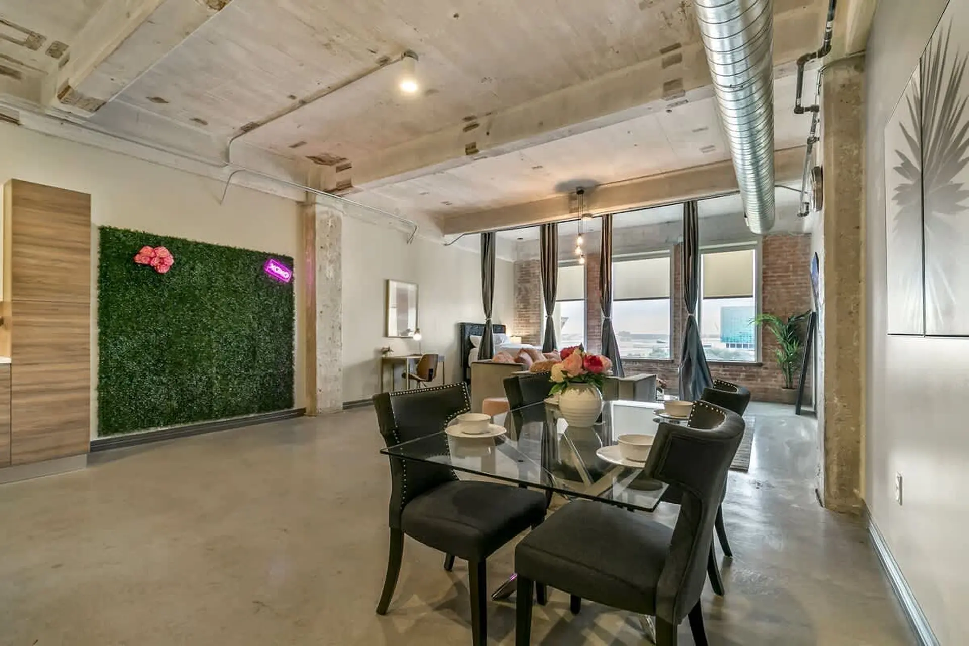 Loft dining area with glass table, plush chairs, and a faux green wall with pink flowers.