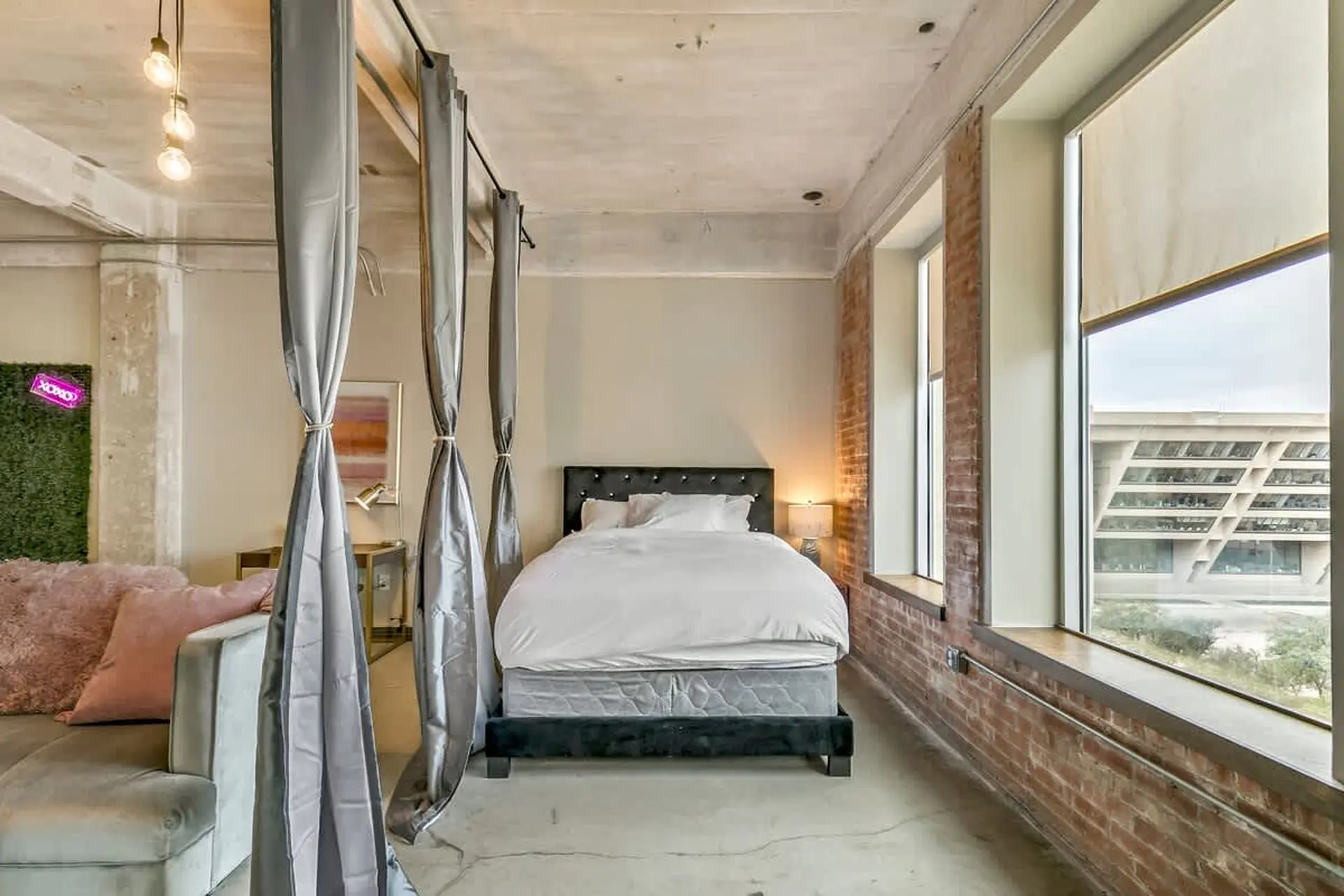 Loft bedroom with exposed brick, industrial lighting, and large windows overlooking a city building.