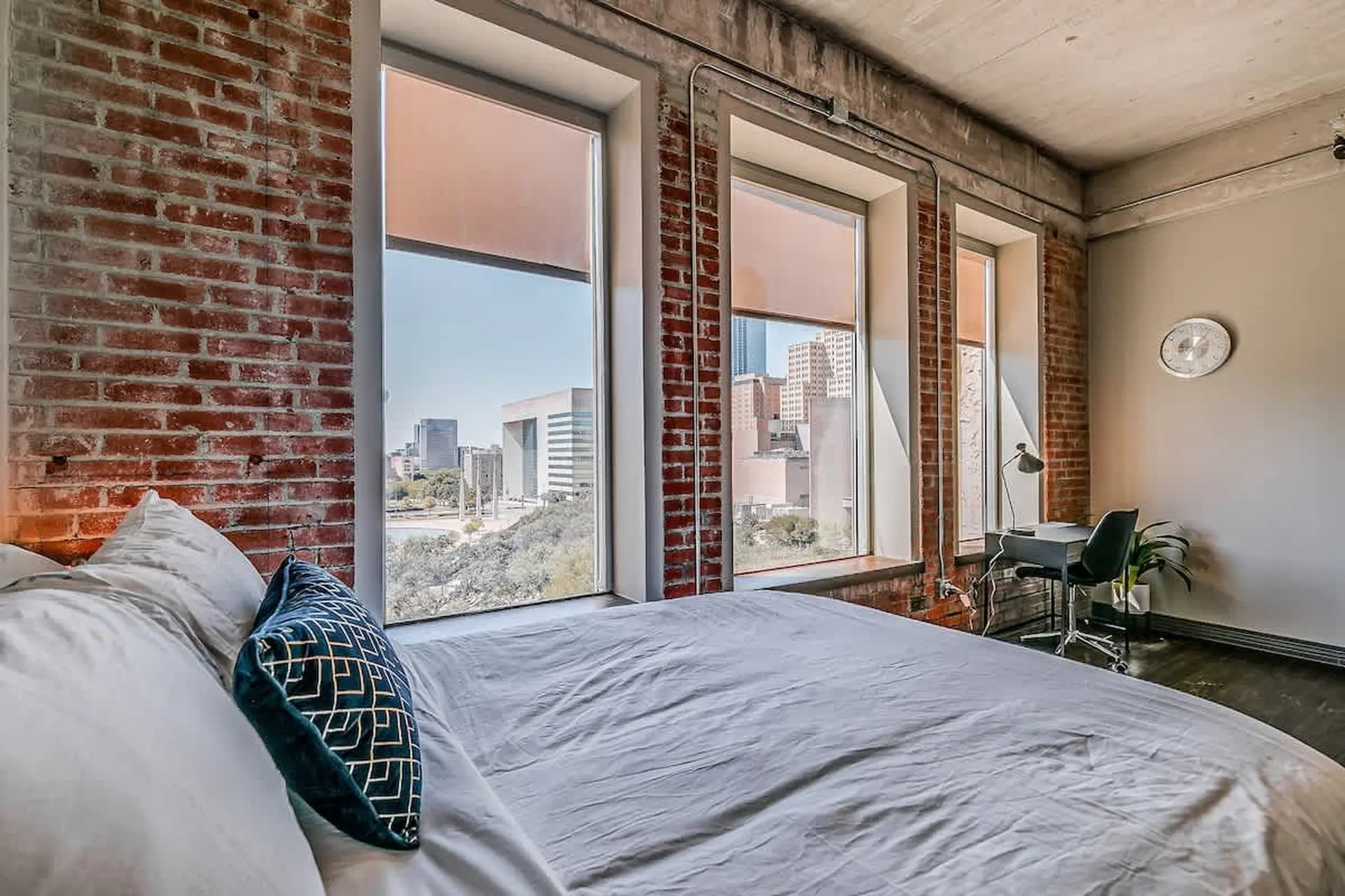 A bed with white linens and blue pillows in front of a large window with a city view.