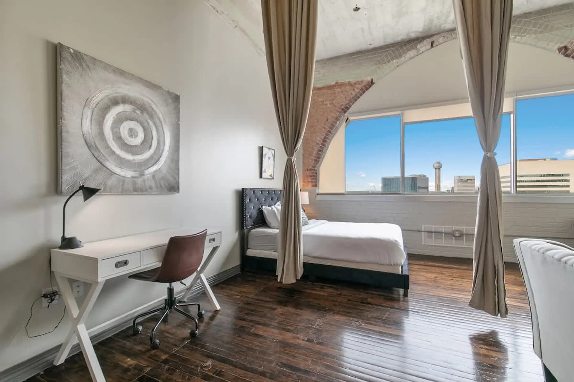 A modern bedroom with a desk, bed, and large windows overlooking a cityscape.