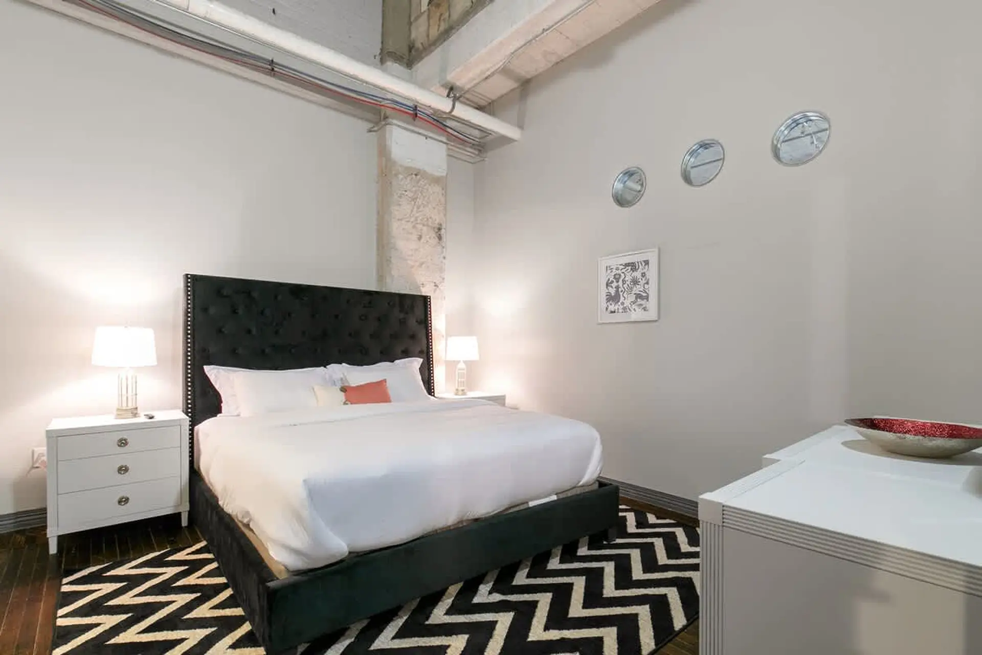 A bedroom with a tufted headboard, white bedding, nightstand with lamp, and a chevron rug.