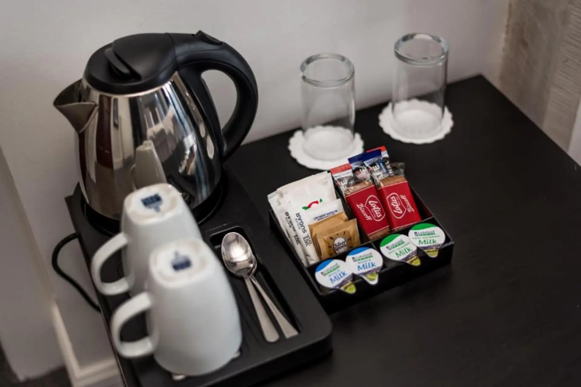 A hotel room coffee and tea setup with electric kettle, cups, sugar, milk, and cookies.