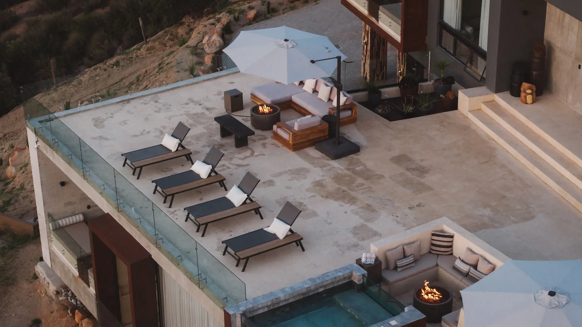 An elevated view of a modern patio with lounge chairs, a sectional sofa with a fire pit, and a swimm
