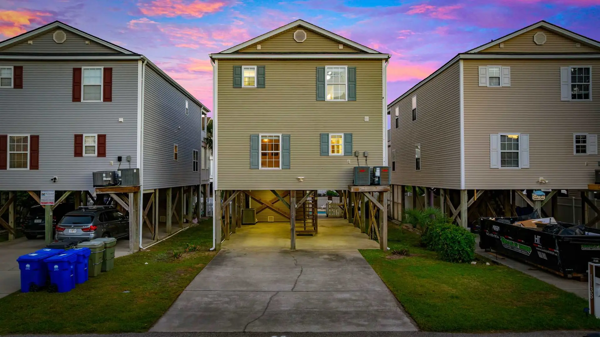 A tan stilt house stands between two gray houses under a colorful twilight sky.