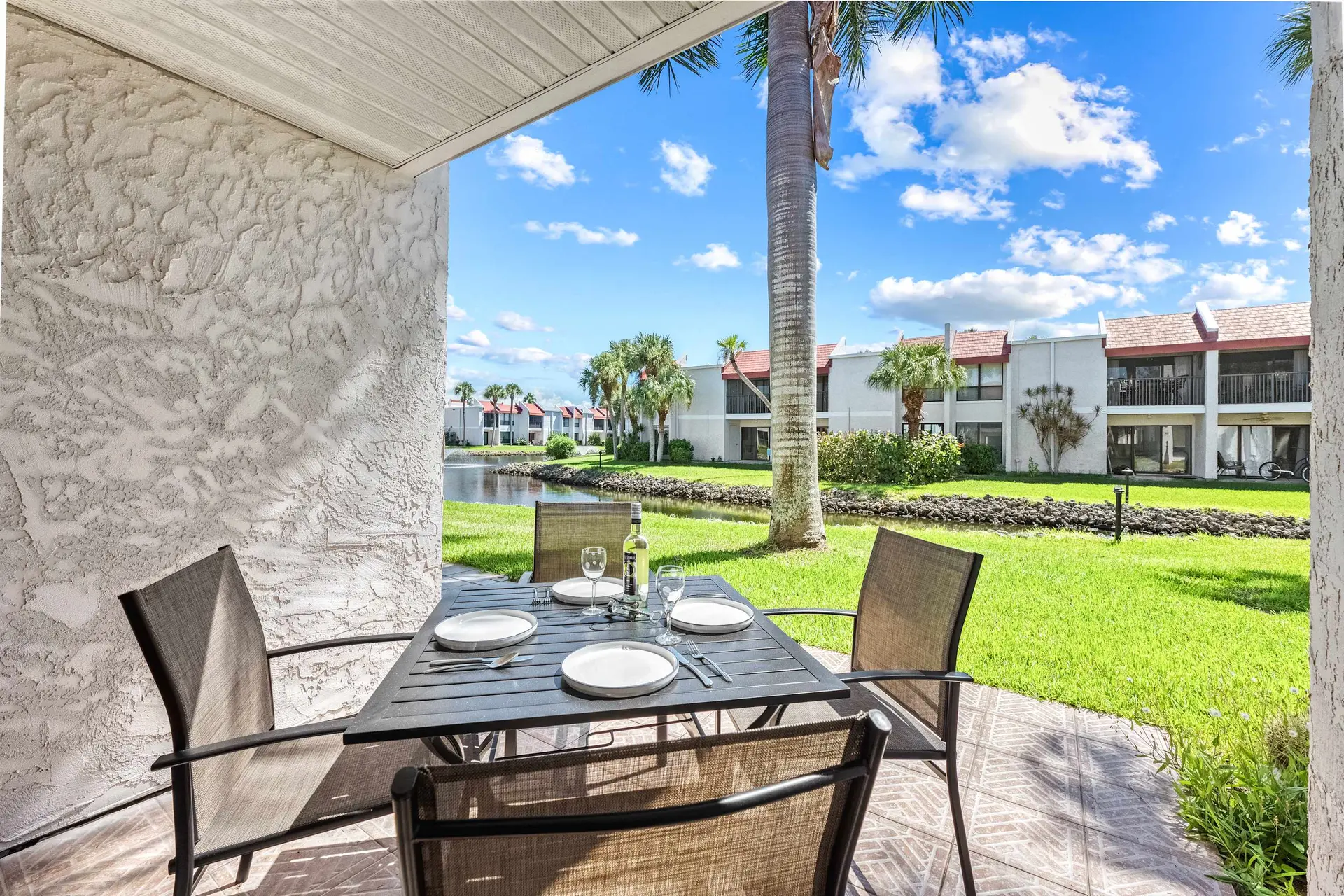 Outdoor dining patio with table set for four, overlooking a pond and condos.