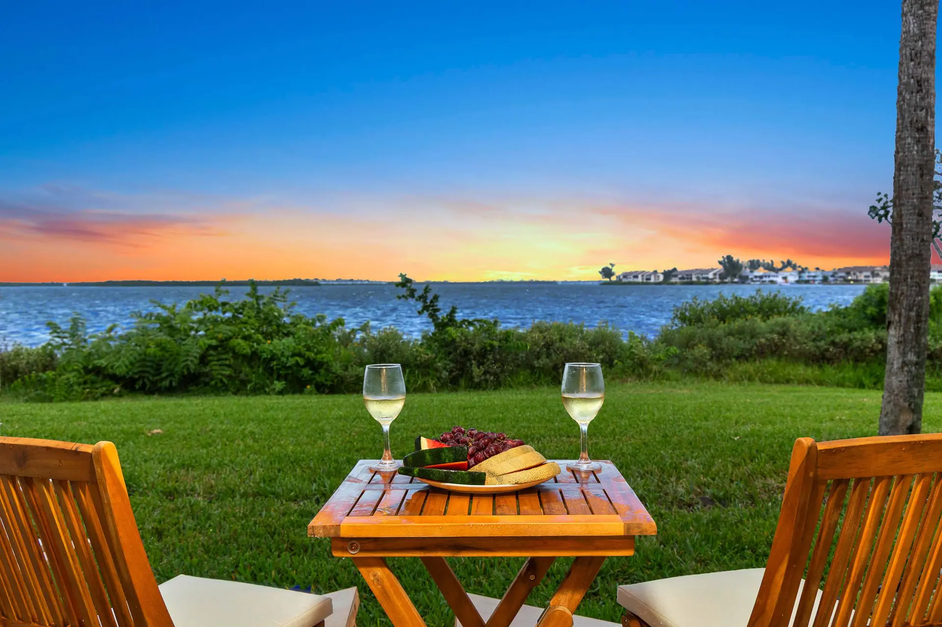 A table with wine and fruit is set for two on a lawn overlooking a lake at sunset.