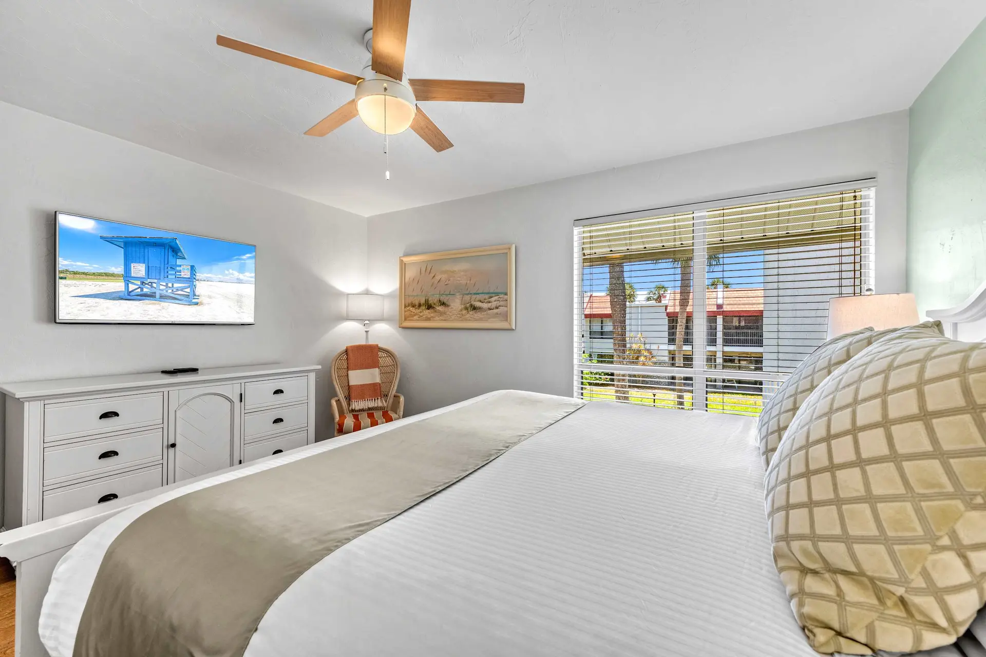 A bedroom with a TV, dresser, chair, painting, ceiling fan, and bed.