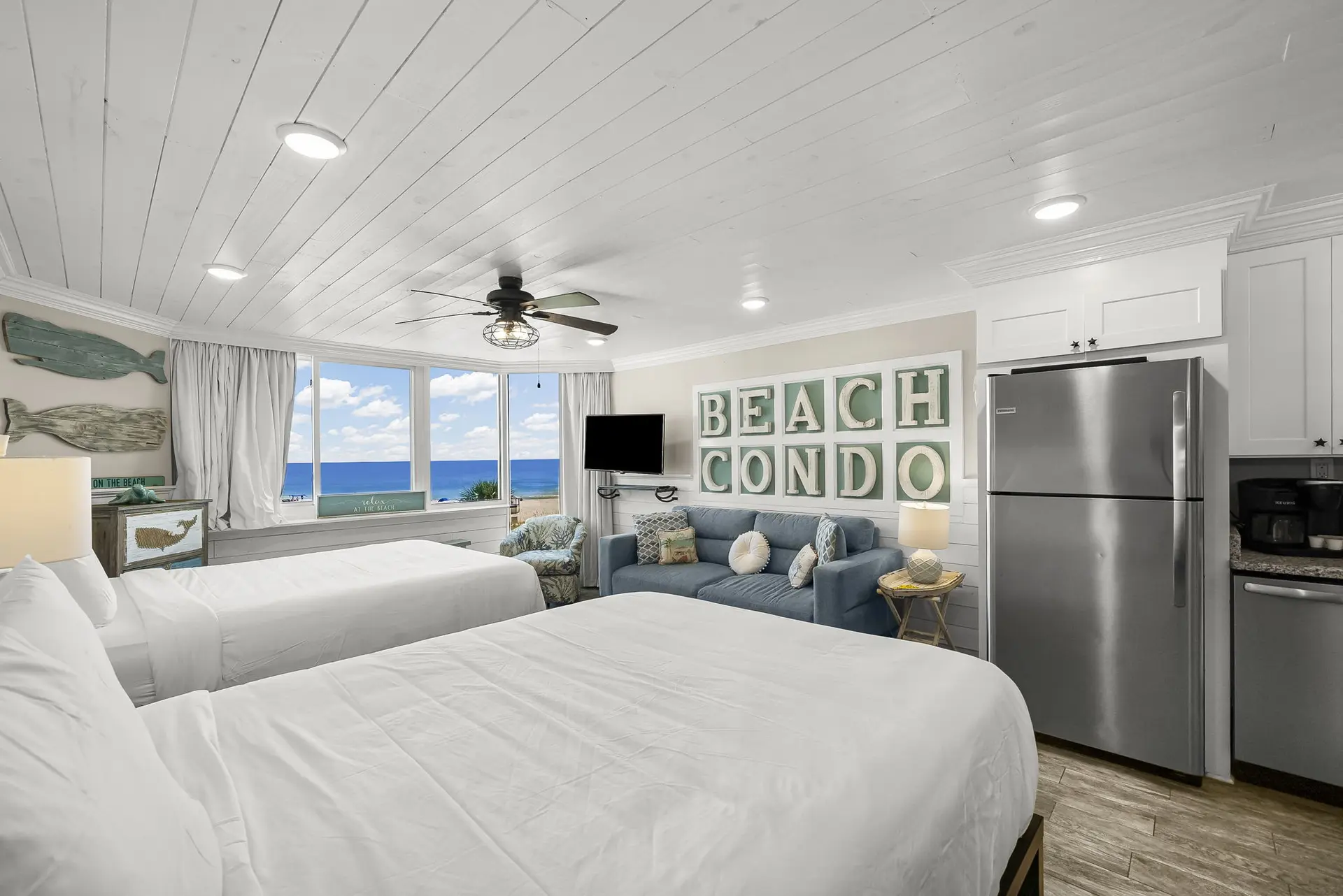A bright, beach-themed hotel room with two beds, a sofa, and a refrigerator.
