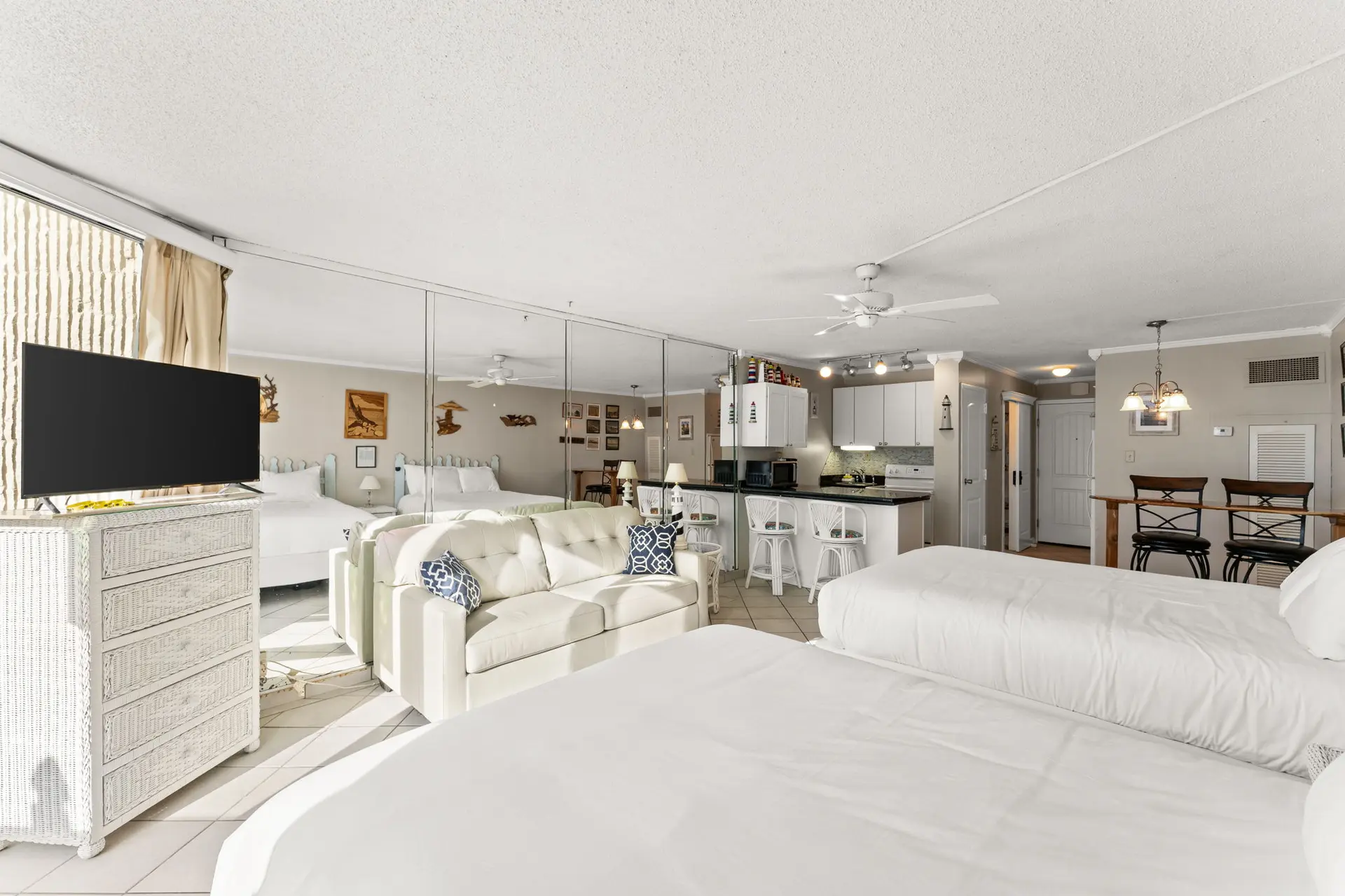 A brightly lit studio apartment with two beds, a sofa, and a kitchen.