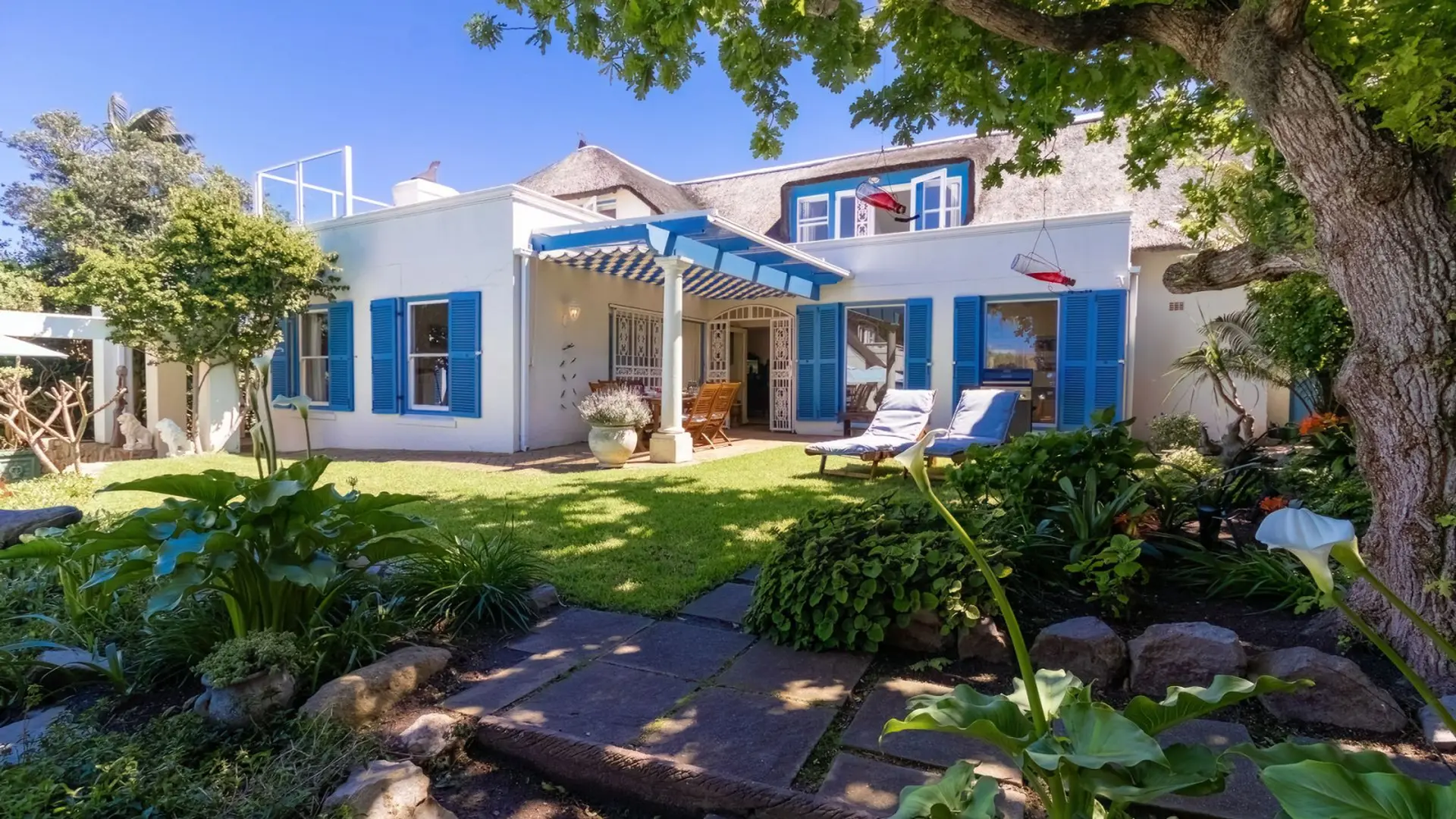A white house with blue shutters and a thatched roof sits on a sunny lawn.