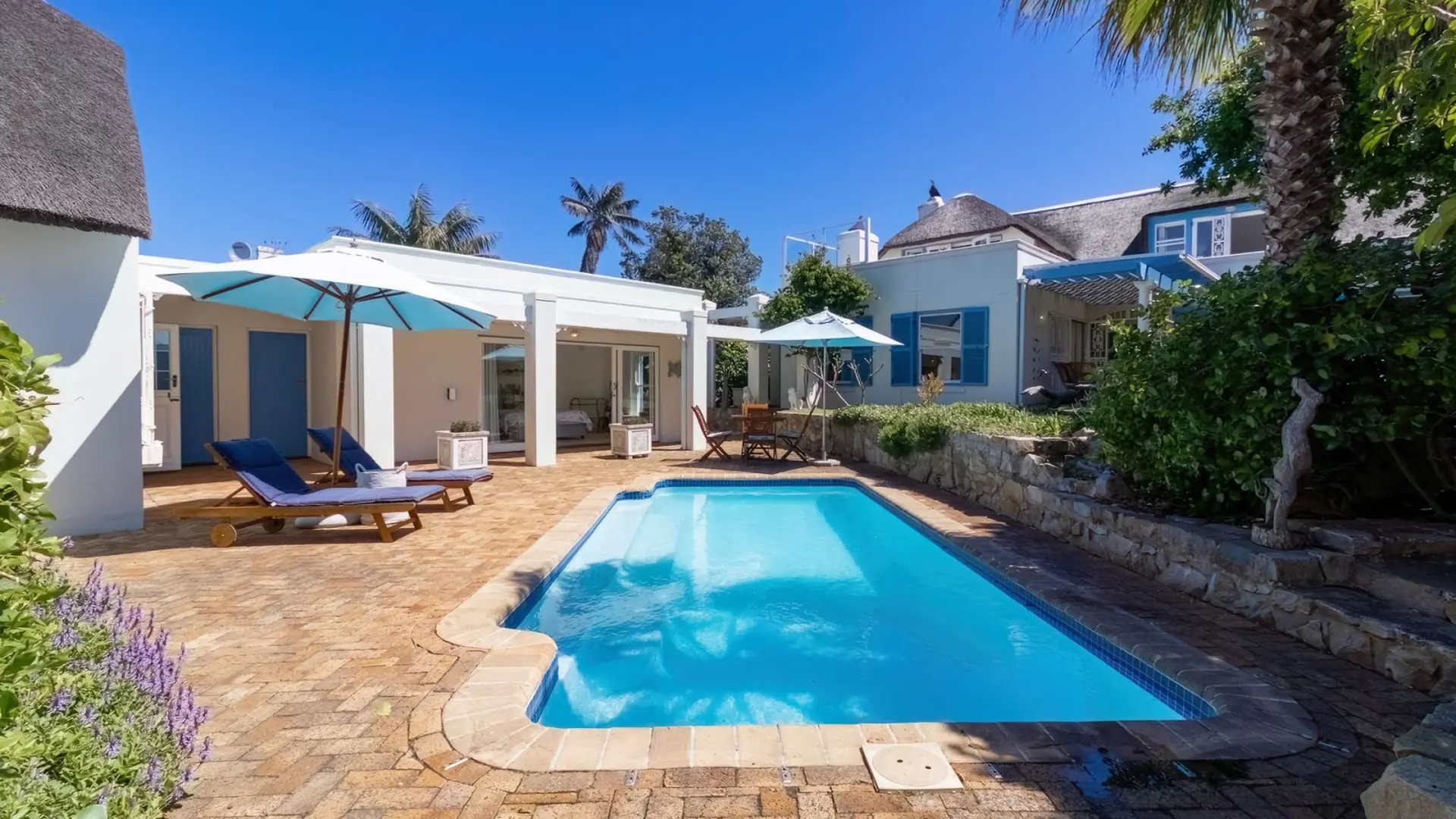 A sunny backyard with a swimming pool, lounge chairs, and blue umbrellas.