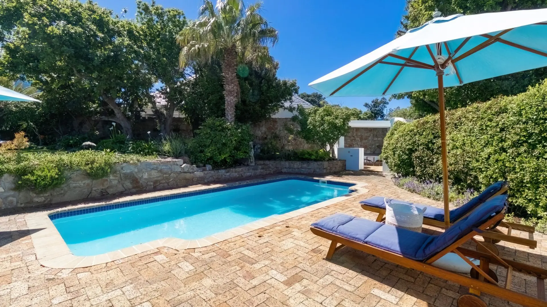 A blue-tiled swimming pool is bordered by a brick patio and lush greenery.