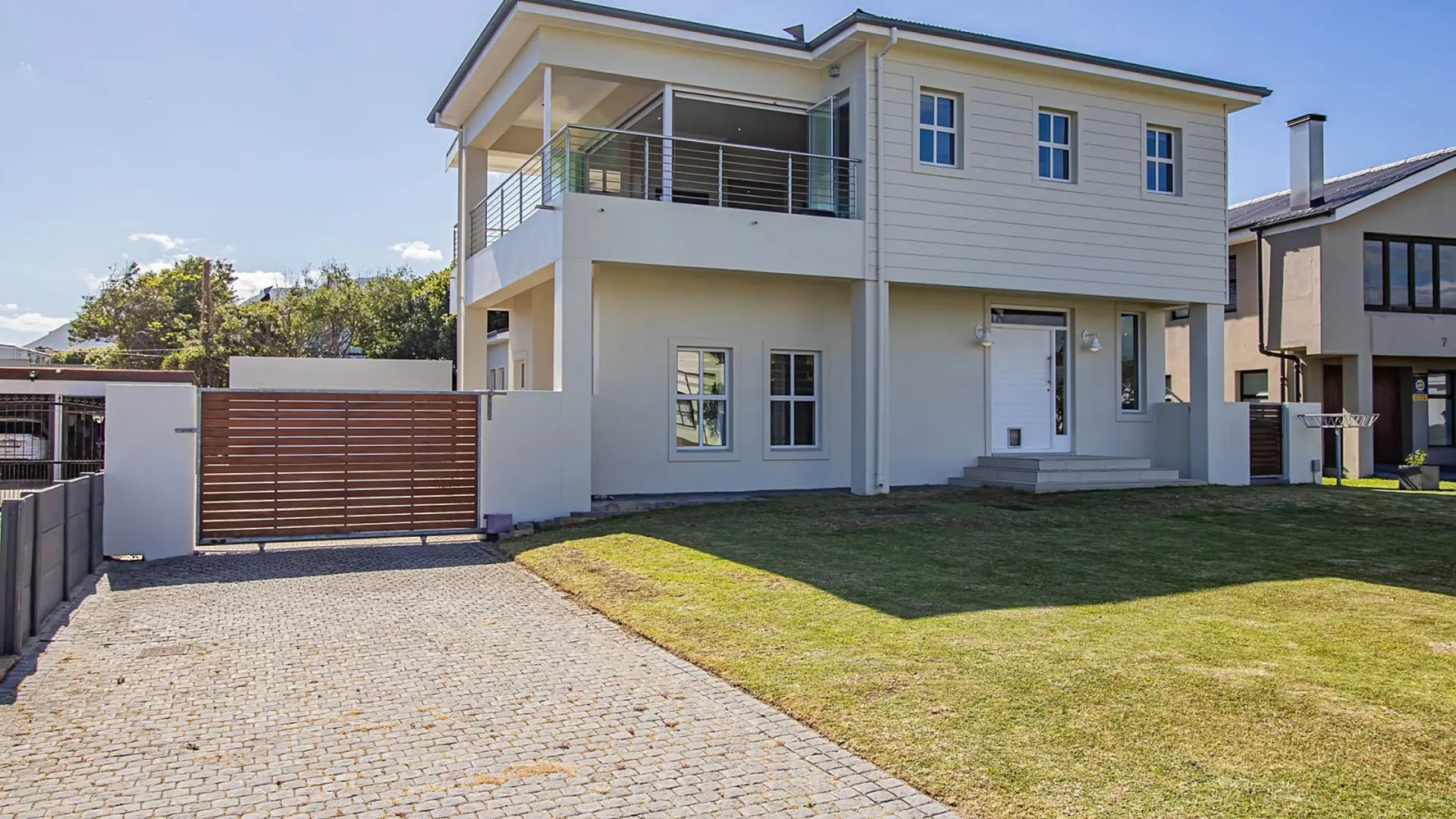 A modern, two-story house with a balcony, driveway, and wooden gate.