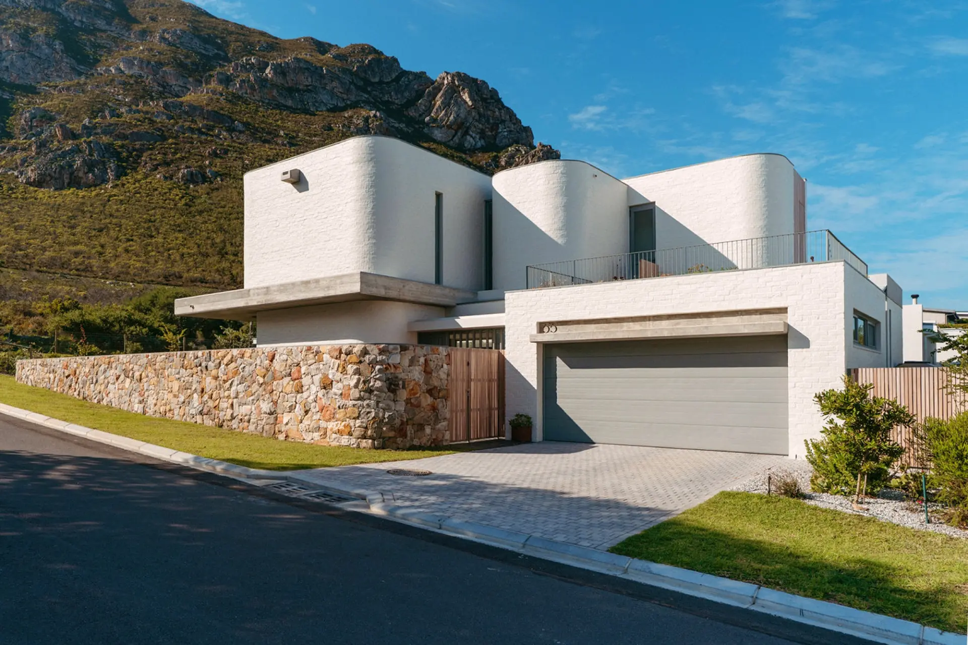 Modern white house with curved walls and a stone wall in front of a mountain.
