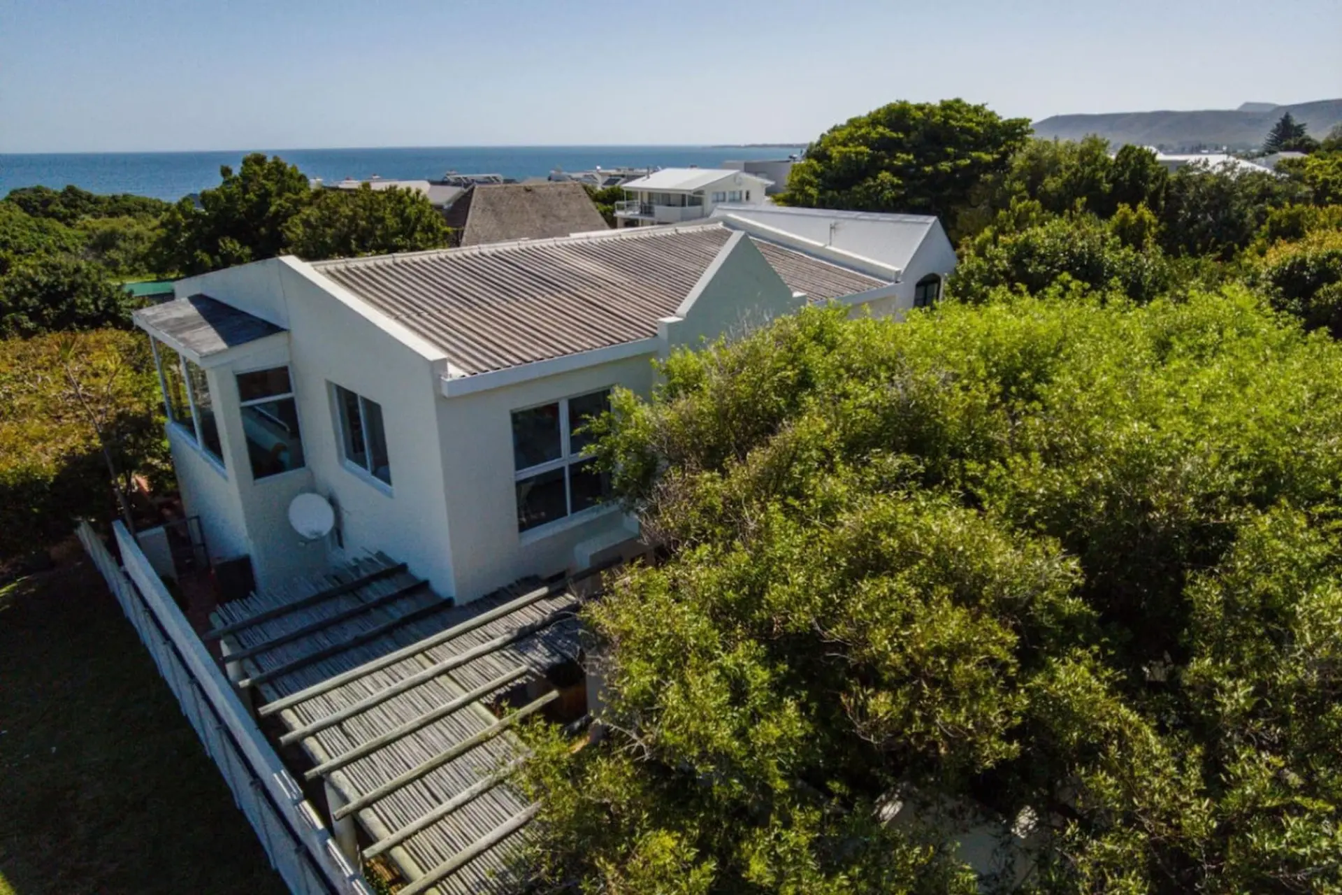 An aerial view of a white house with a grey roof, surrounded by lush greenery and the ocean in the b