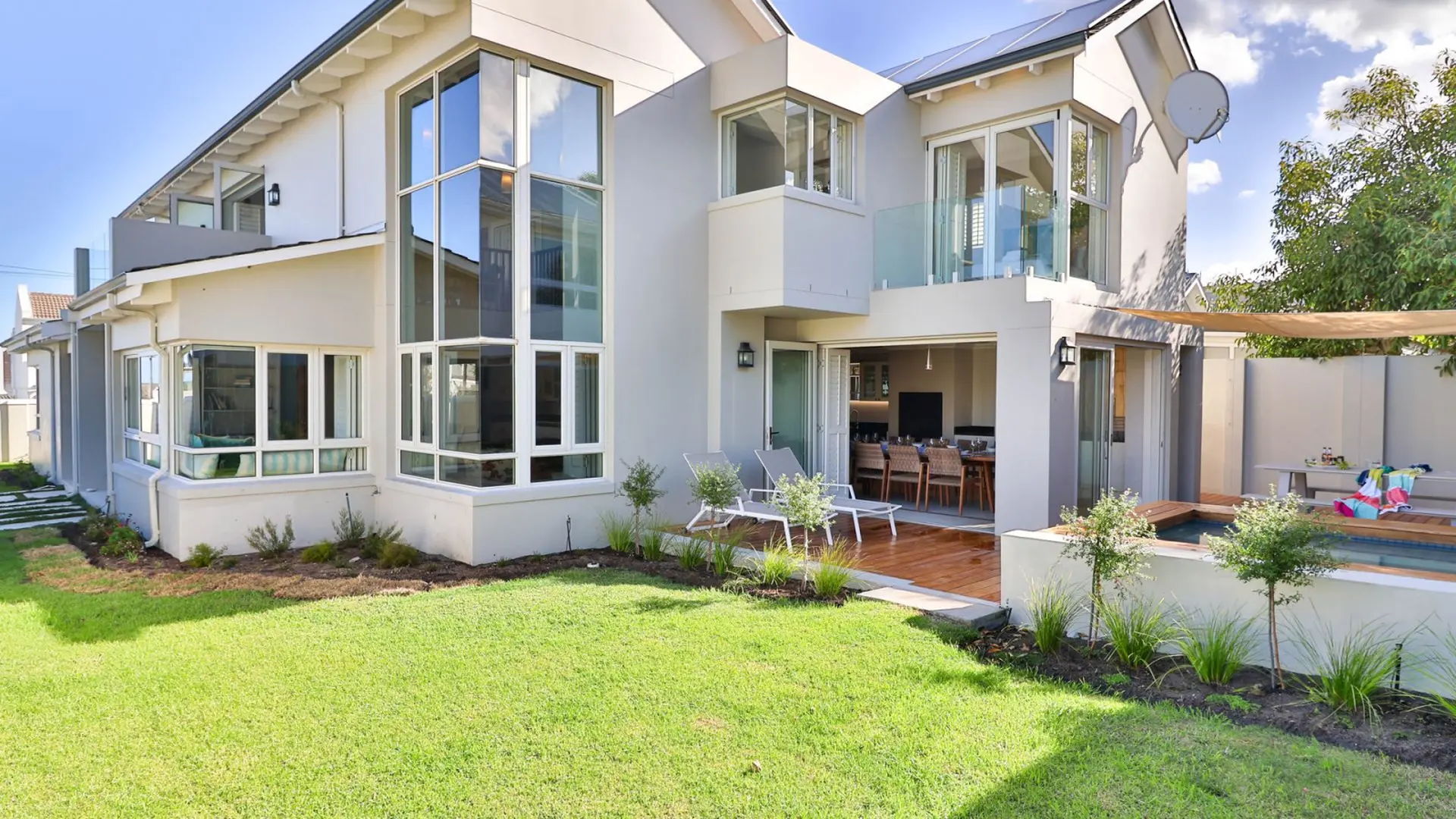 A modern white house with large windows, a wooden deck, and a swimming pool.