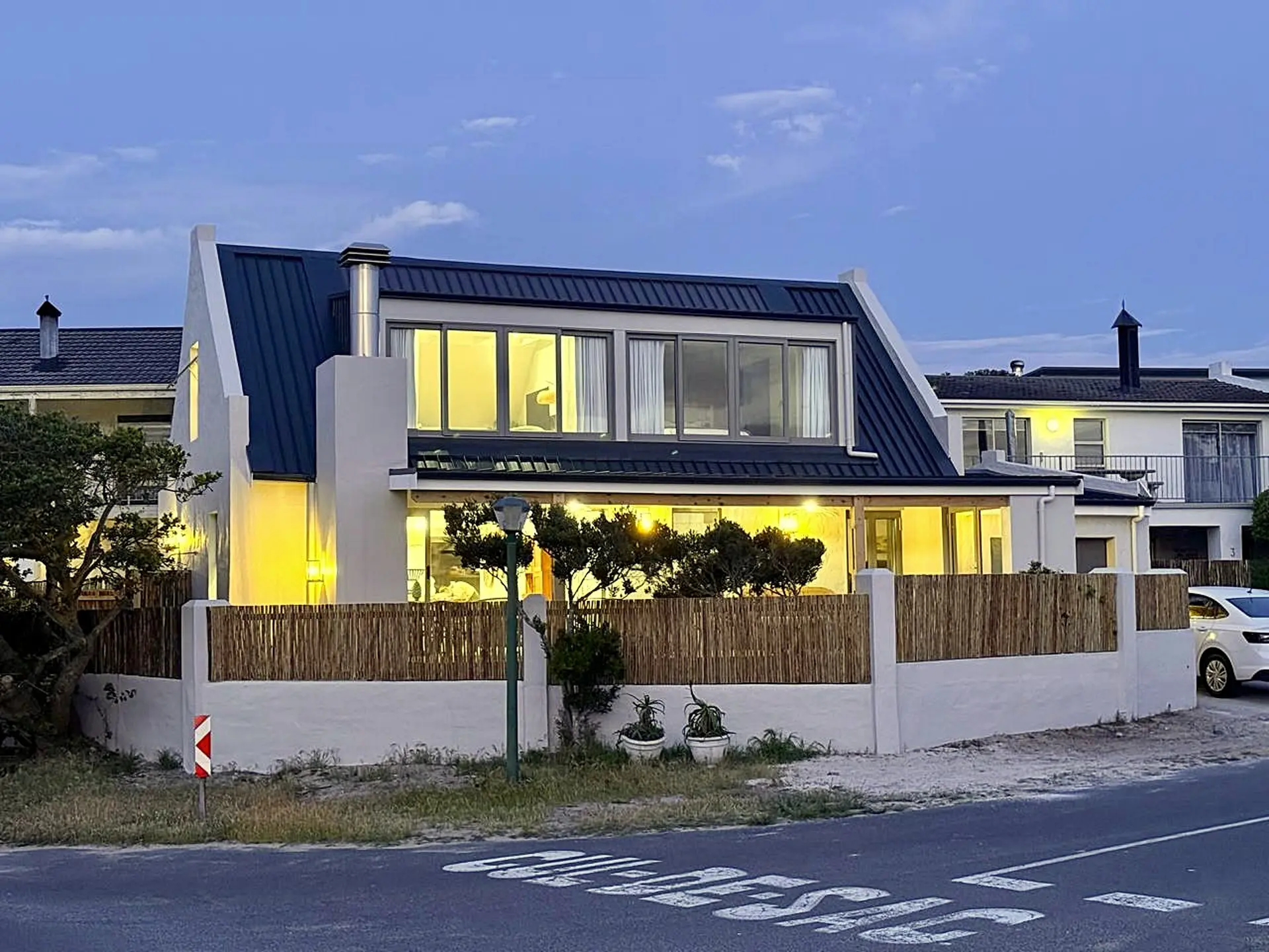 A modern white house with a dark metal roof, lit up at dusk, with a bamboo fence.