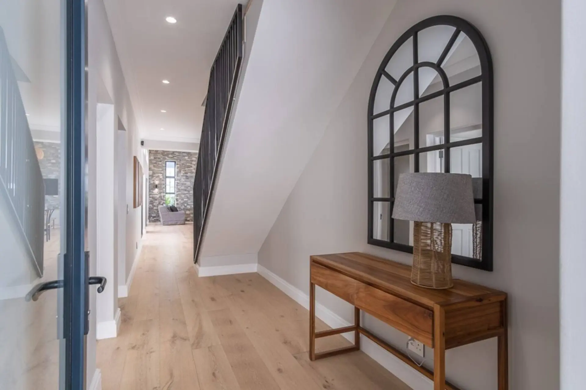 A hallway with a wooden floor, a black staircase railing, and a wooden console table with a lamp.