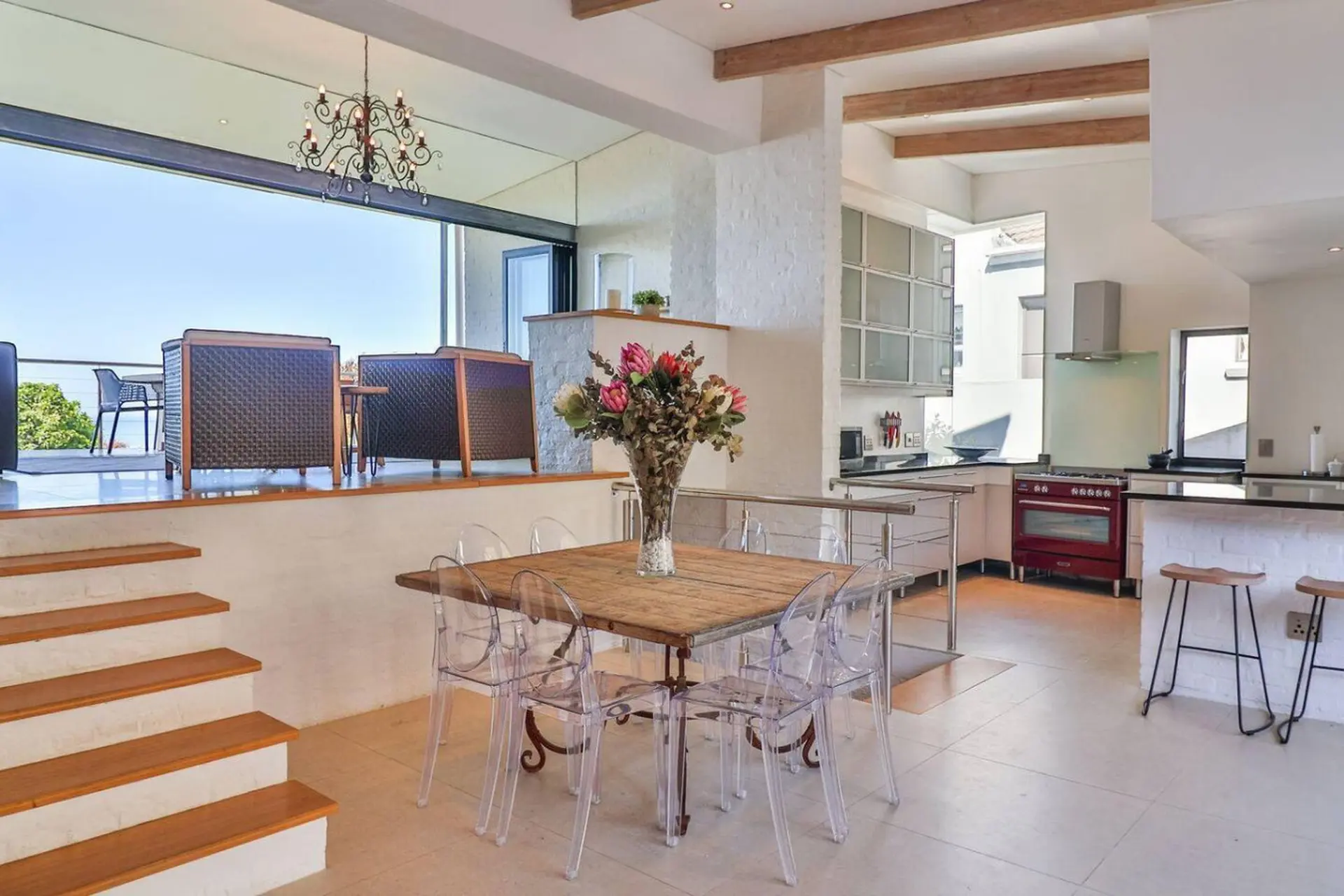 A bright, modern dining and kitchen area with a red stove, clear chairs, and a vase of flowers.
