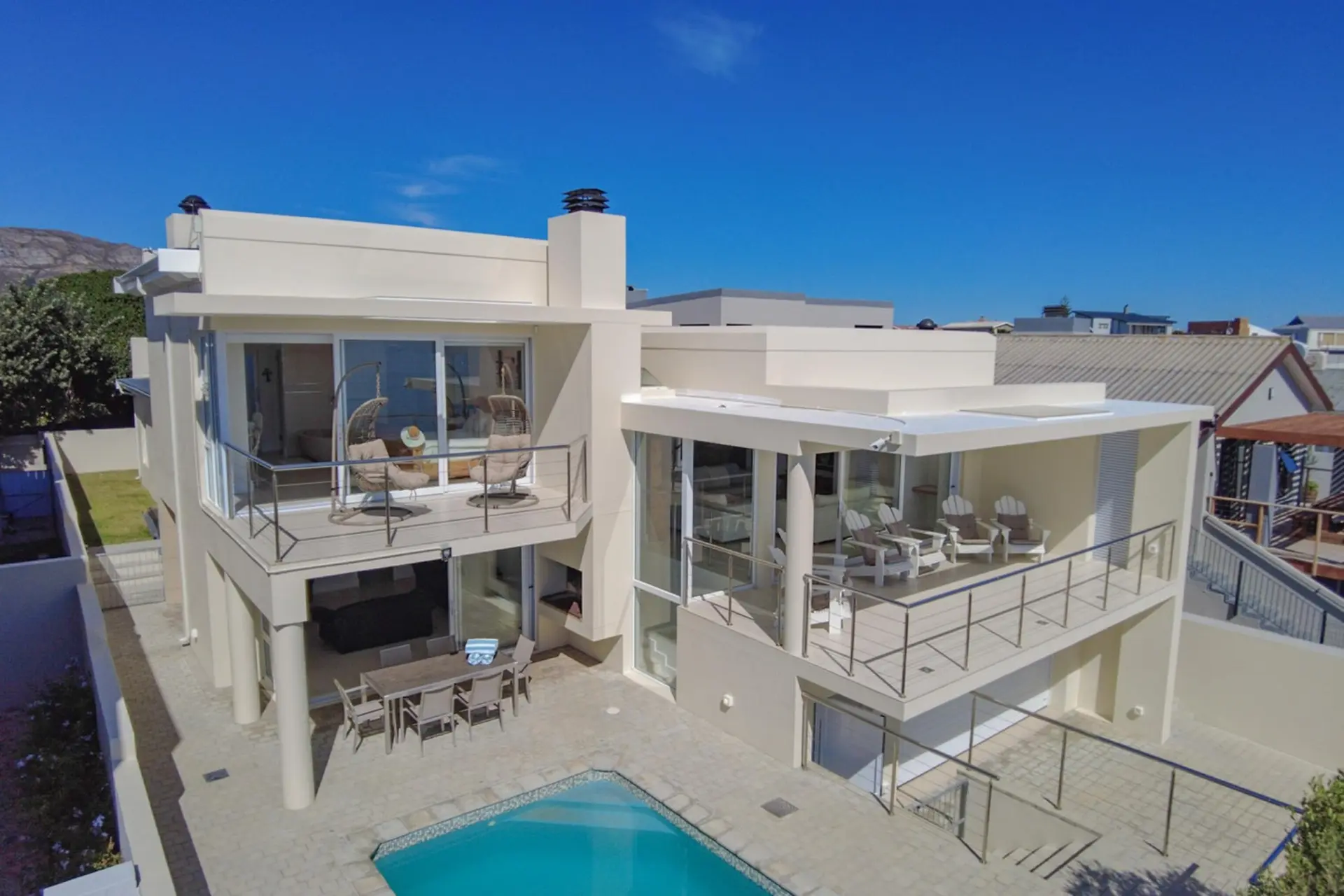 Modern cream-colored house with a pool, balconies, and outdoor furniture on a sunny day.