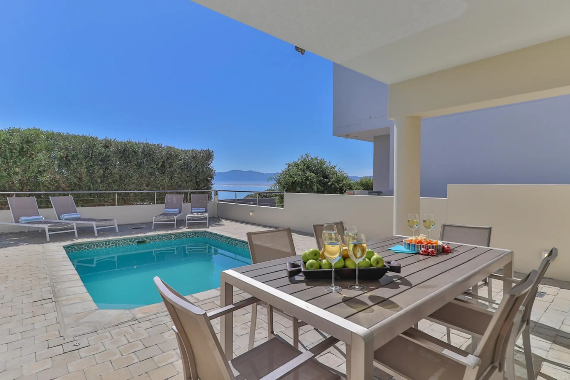 A patio with a table and chairs overlooks a pool and the ocean.