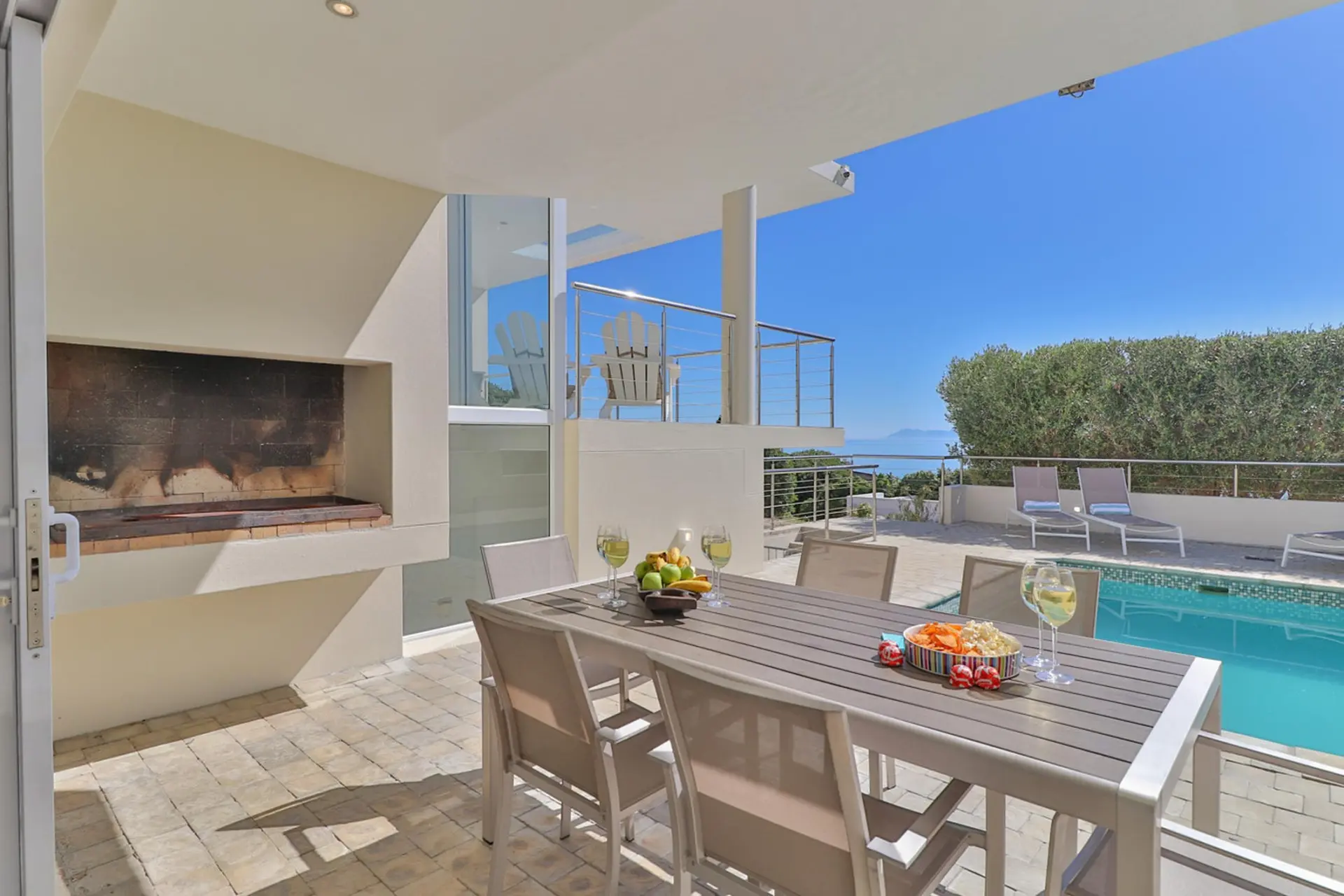 Outdoor dining area with a table and chairs by a pool and a grill, overlooking the ocean.