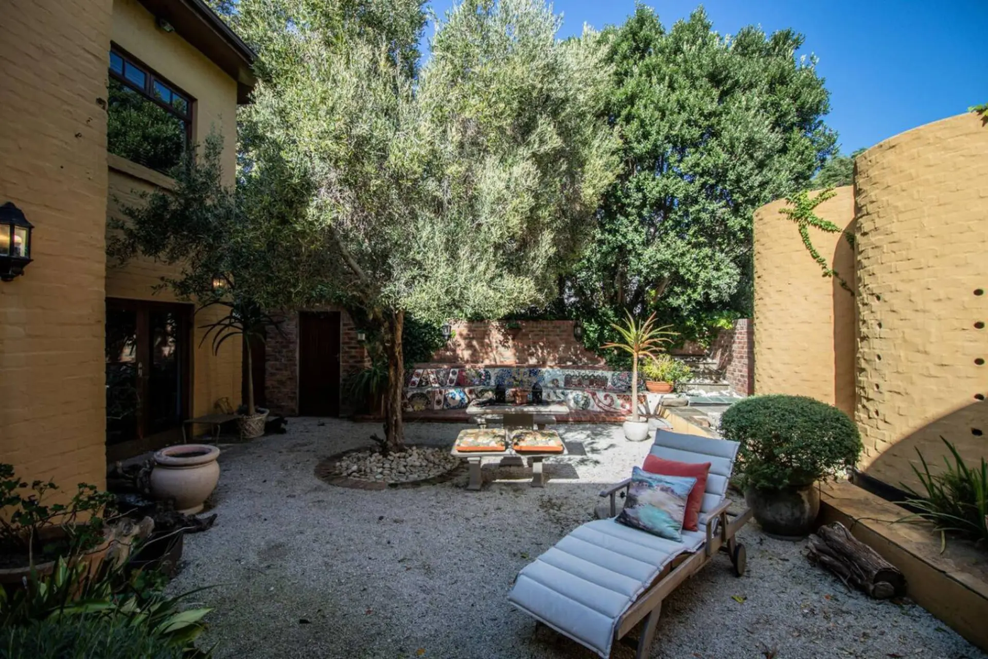 A sunny courtyard with a lounge chair, potted plants, a mosaic bench, and an olive tree.