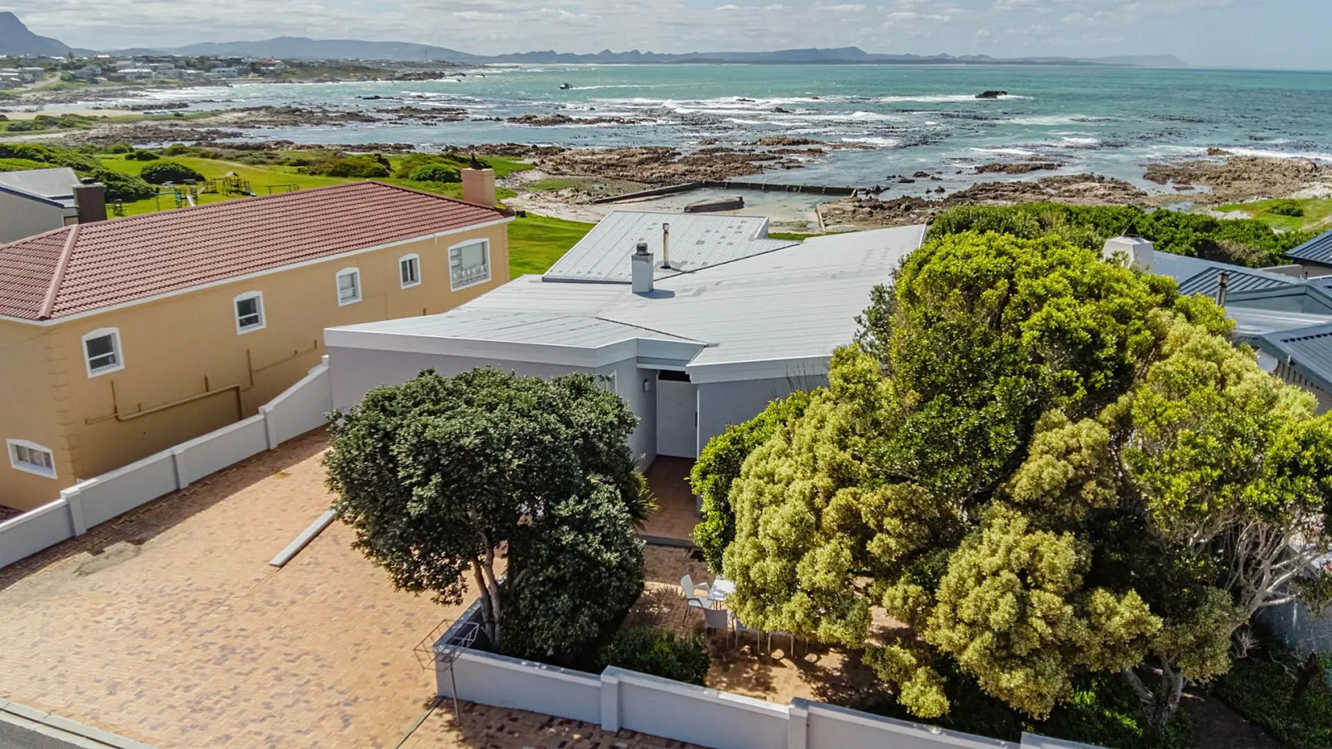 Aerial view of a house with a gravel driveway and trees in front of the ocean.
