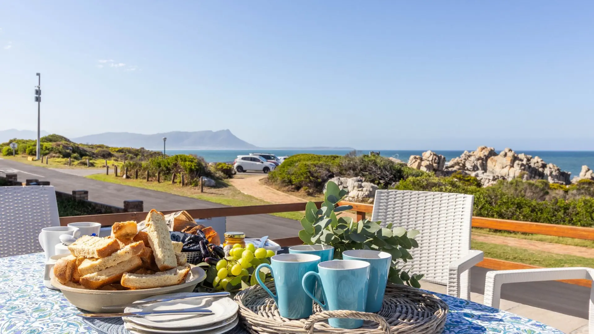 Breakfast spread with bread, fruit, and coffee cups on a table with a sea view.
