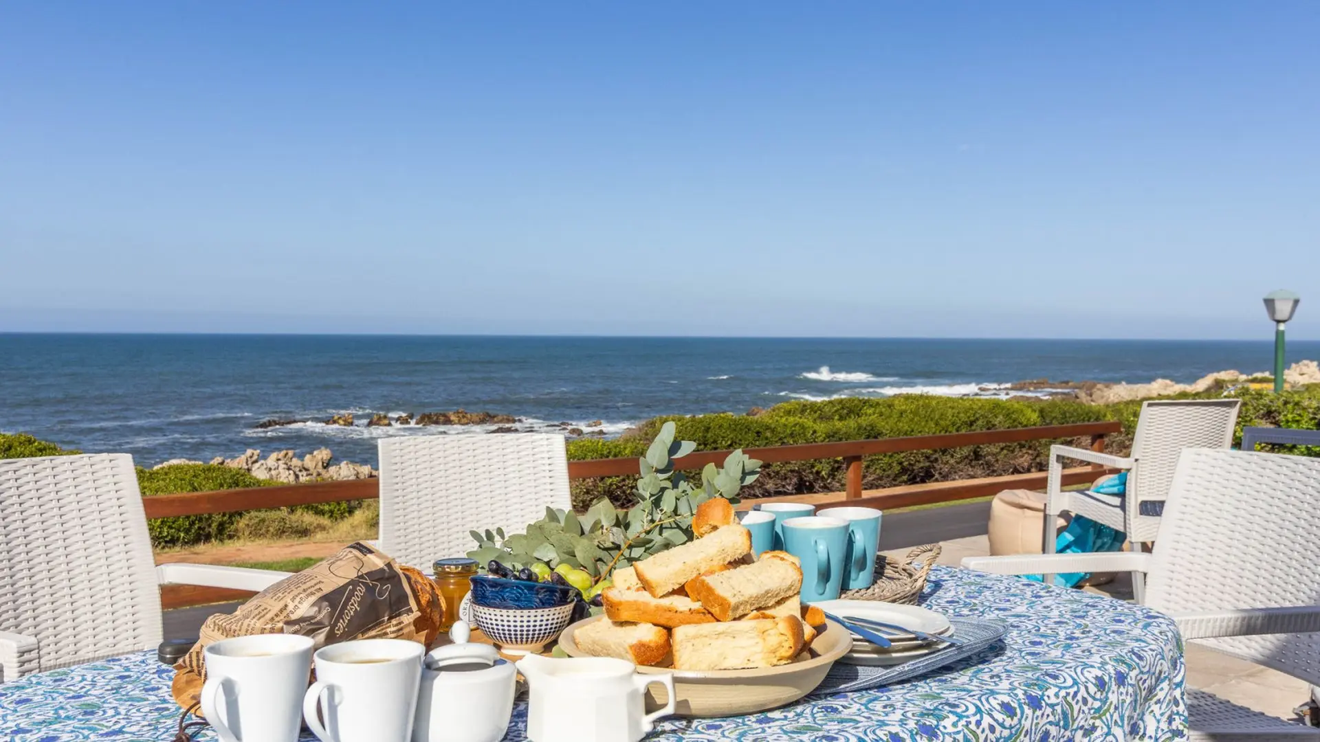A table set for breakfast overlooks the ocean, with white chairs and blue cups.