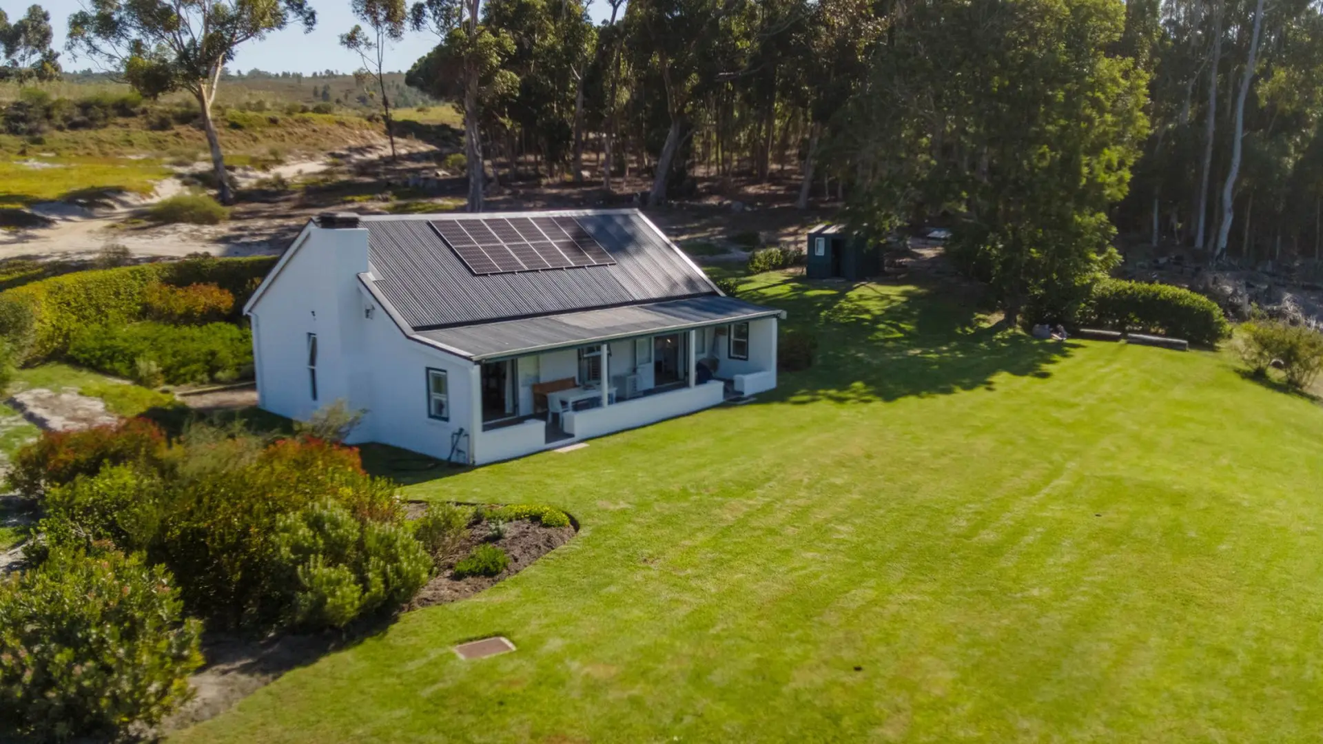 An aerial view of a white cottage with solar panels on the roof, surrounded by green grass and trees