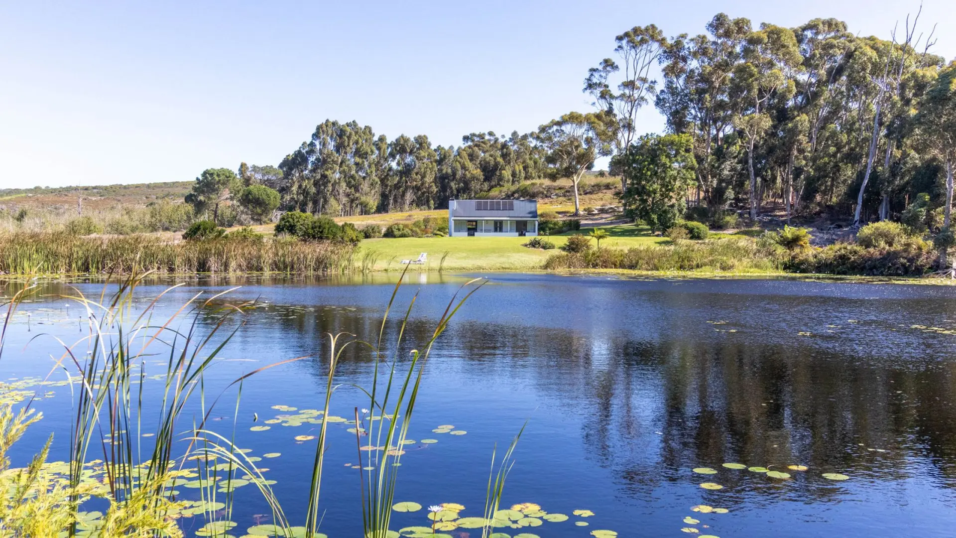 A modern house with solar panels sits on a grassy hill overlooking a tranquil lake.