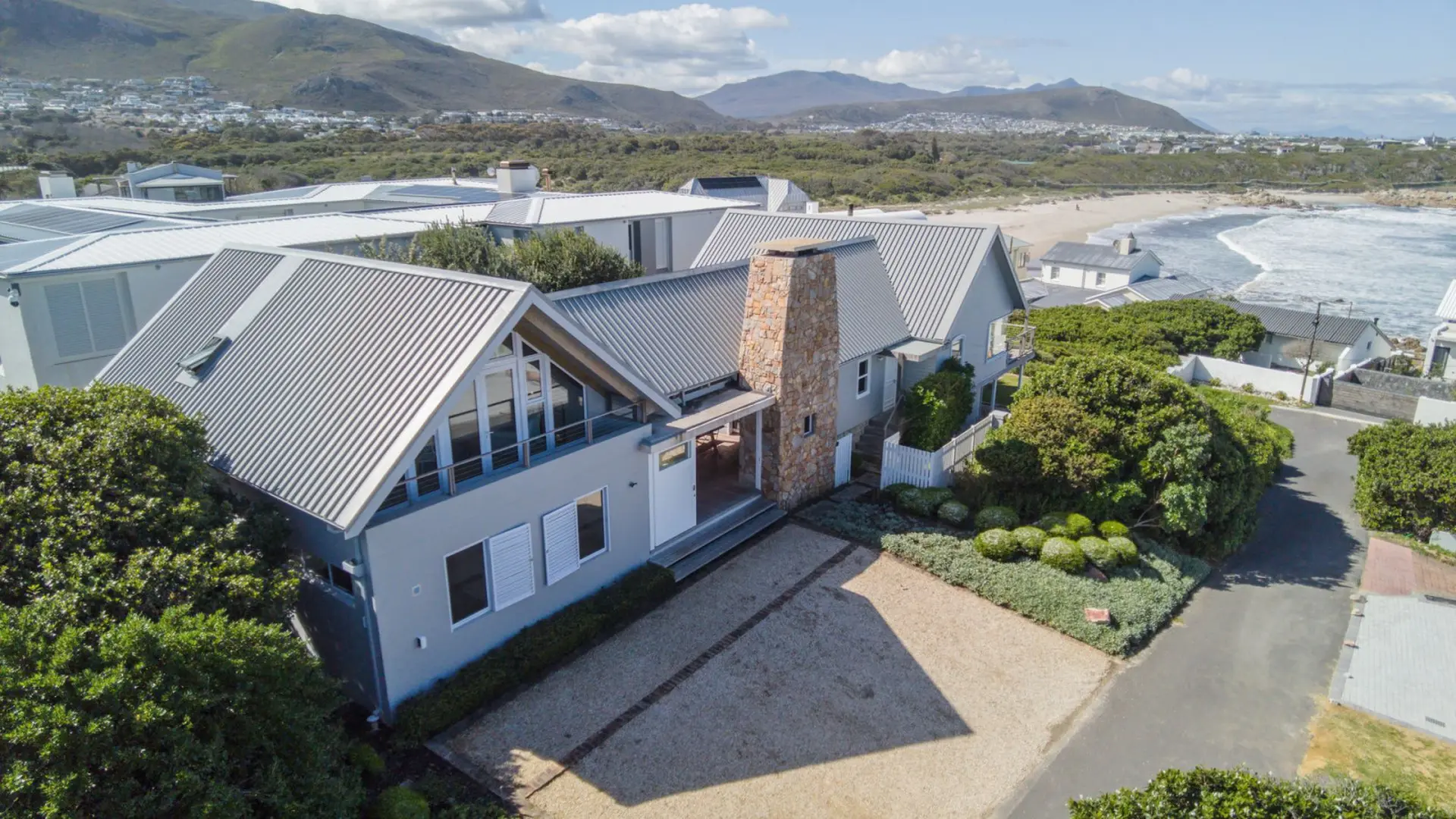 Modern grey house with a stone chimney near a sandy beach and mountains.
