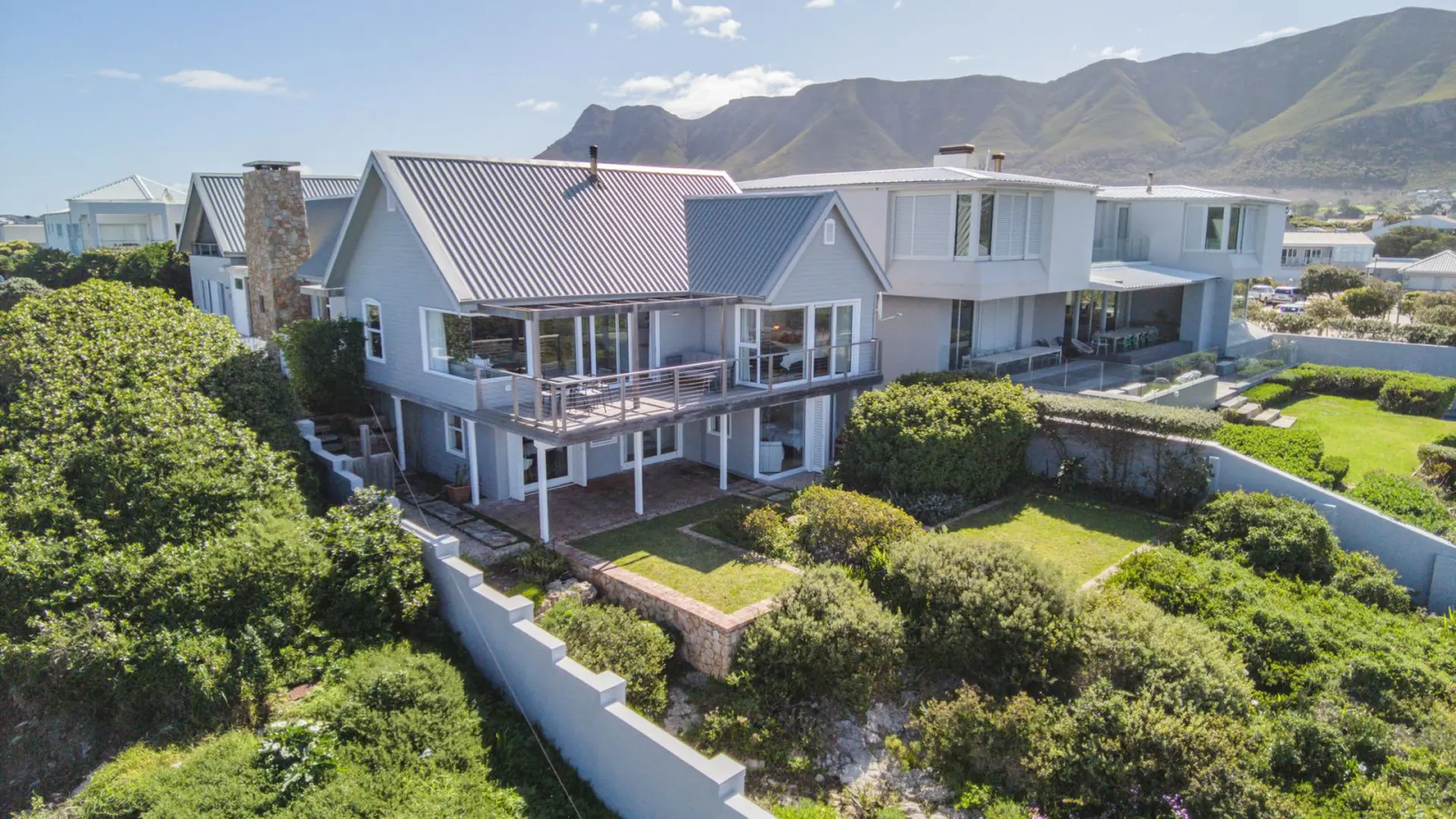 A modern grey house with multiple levels and large windows overlooks a green garden and mountains.
