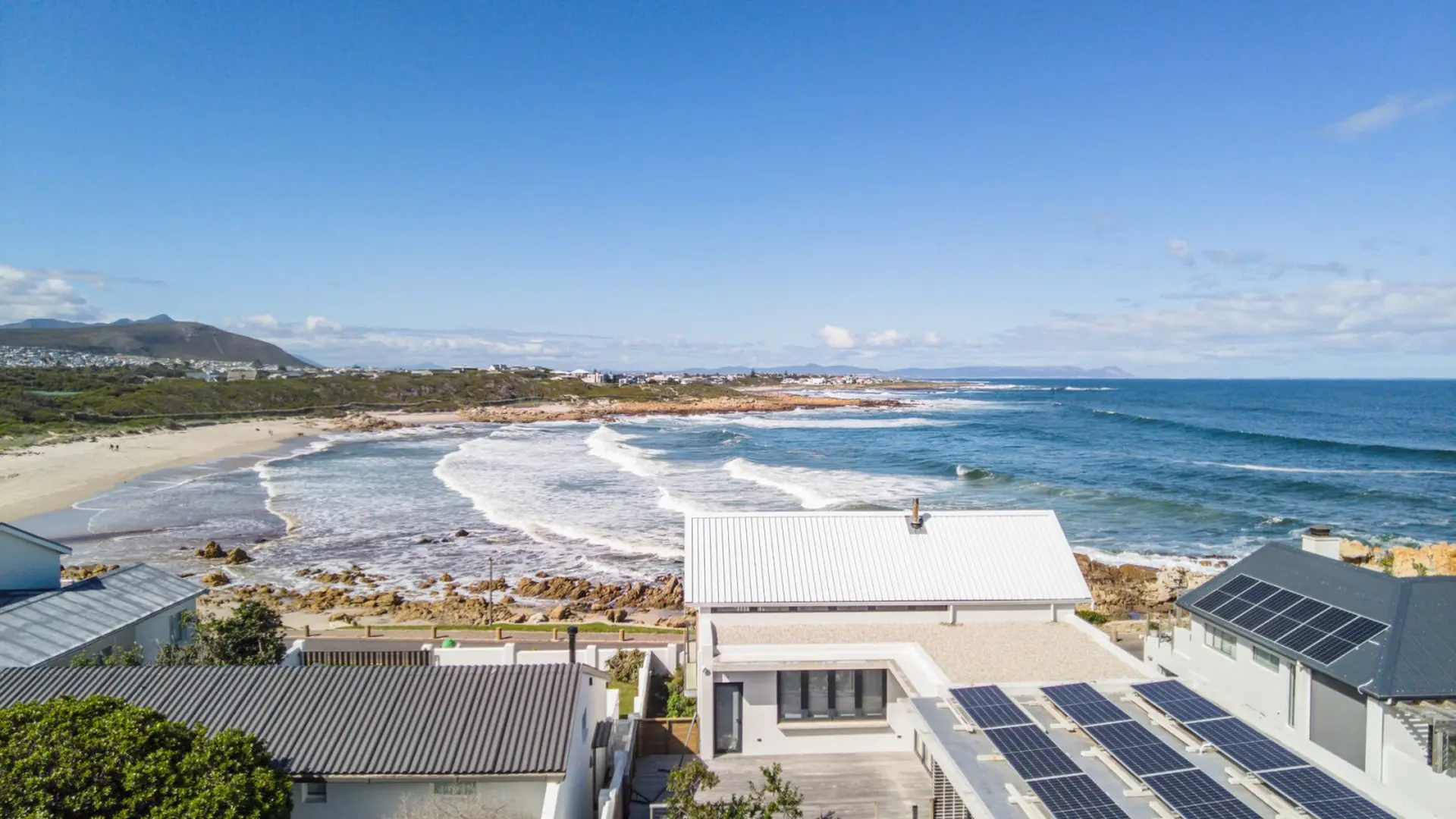 Modern homes with solar panels on a rocky coastline with a sandy beach and blue ocean.