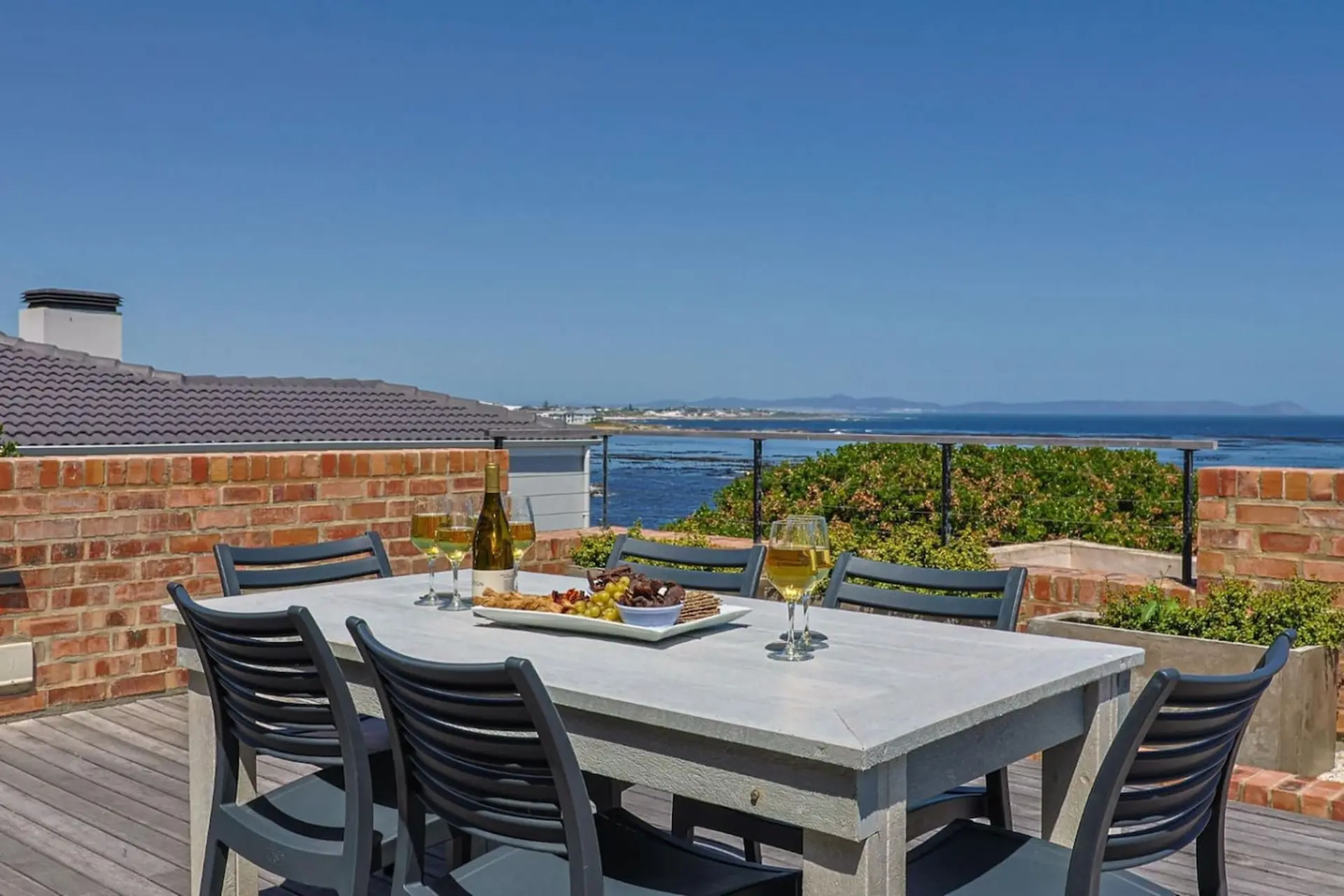 A grey dining table with chairs and a charcuterie board sits on a wooden deck with a view of the oce
