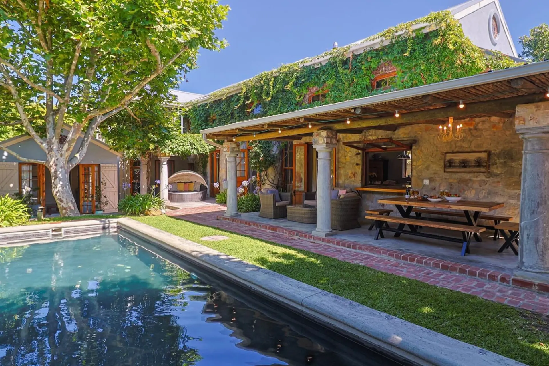 A luxurious poolside patio with a stone house covered in ivy and an outdoor dining area.