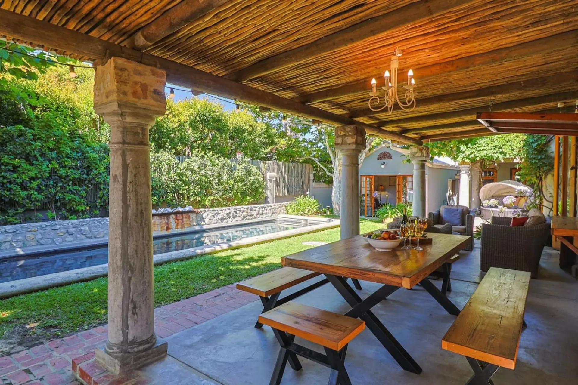 Outdoor dining area with wooden table and benches, pool, and lush greenery.