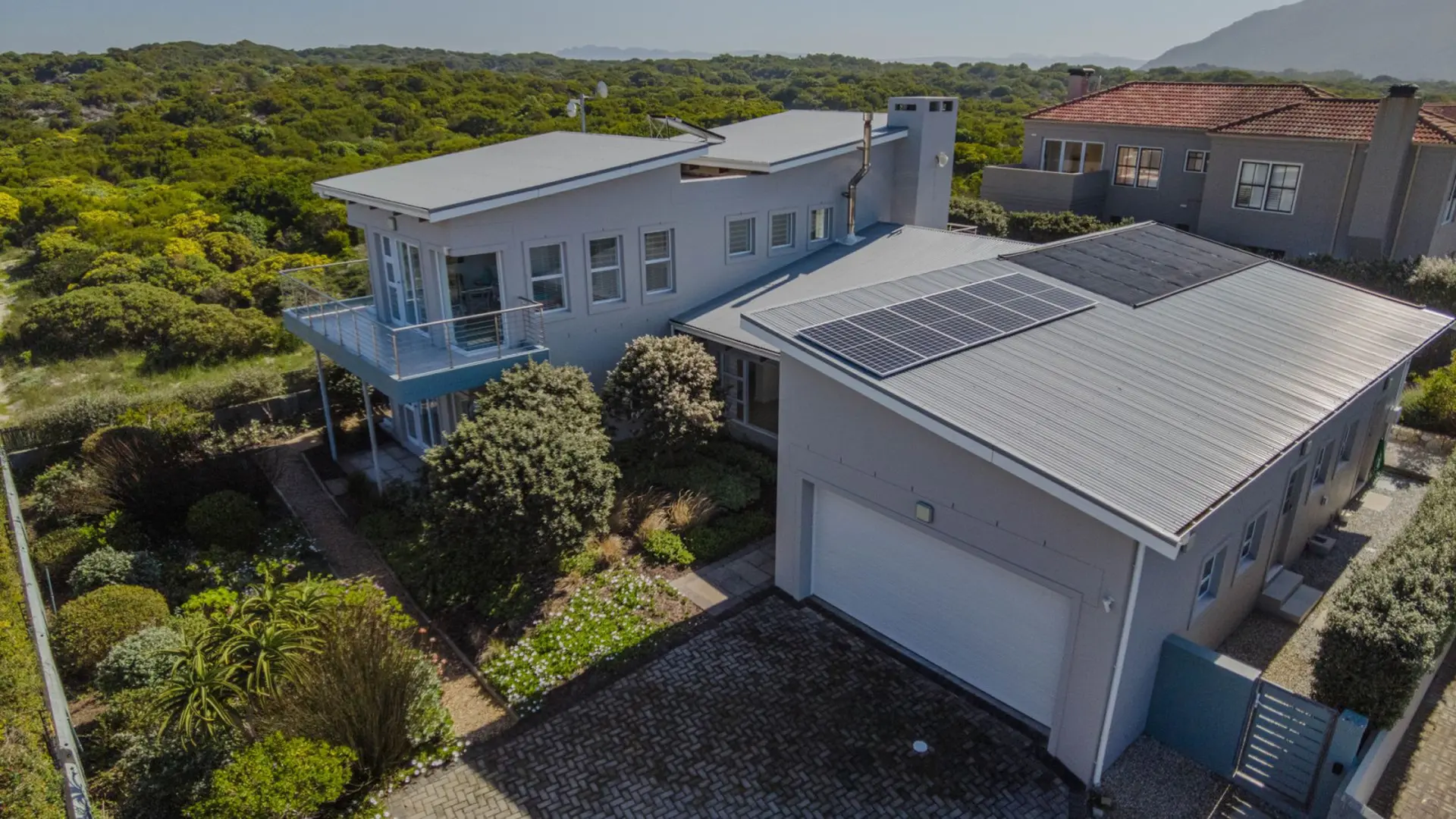 Modern house with solar panels and a balcony, surrounded by lush green trees and shrubs.